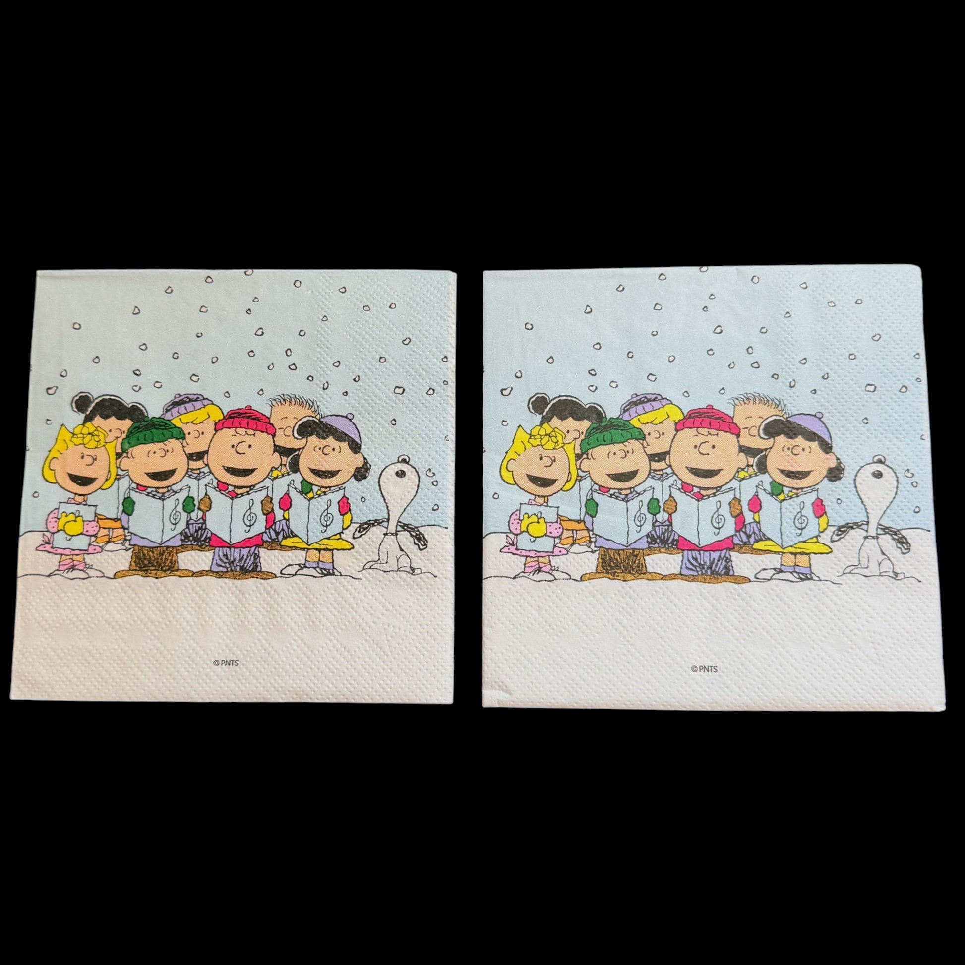 Cocktail size decoupage napkin featuring the Peanuts gang including Charlie Brown Snoopy Lucy Sally and friends caroling together in falling snow on a soft blue winter background set of 2