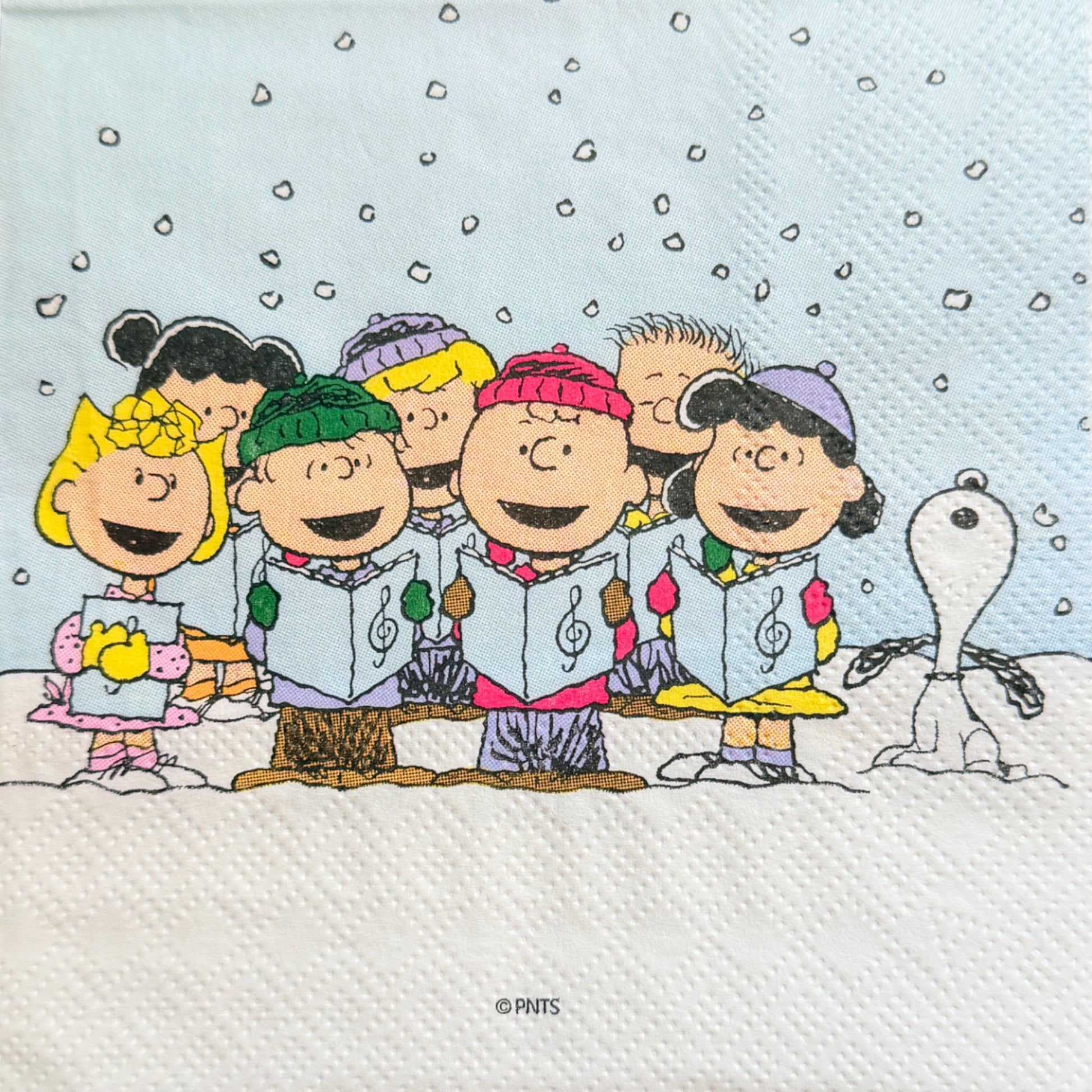 Cocktail size decoupage napkin featuring the Peanuts gang including Charlie Brown Snoopy Lucy Sally and friends caroling together in falling snow on a soft blue winter background set of 2