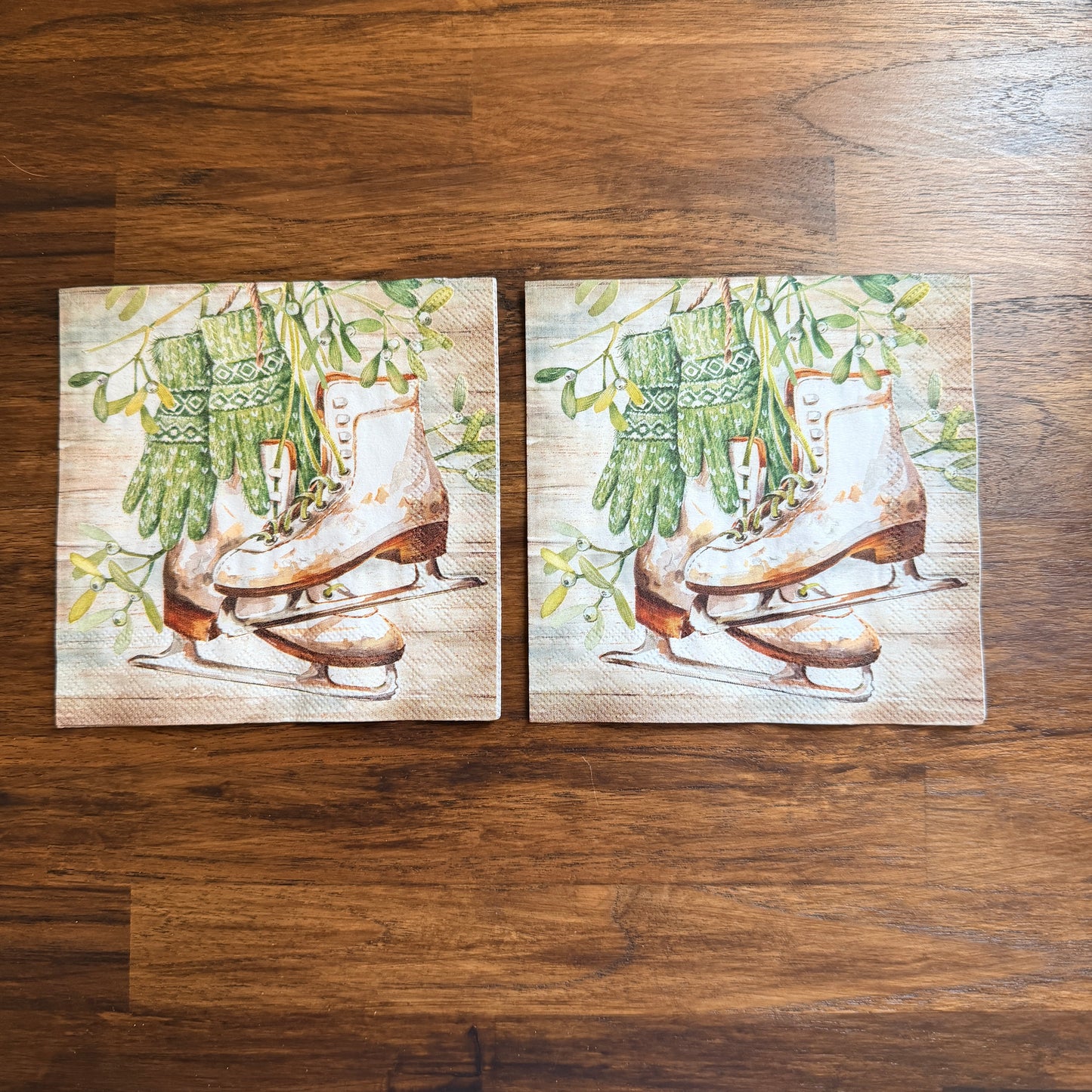 Cocktail size decoupage napkin featuring vintage white leather ice skates with green fair isle mittens hanging from wild mistletoe sprigs on a warm wood grain background set of 2