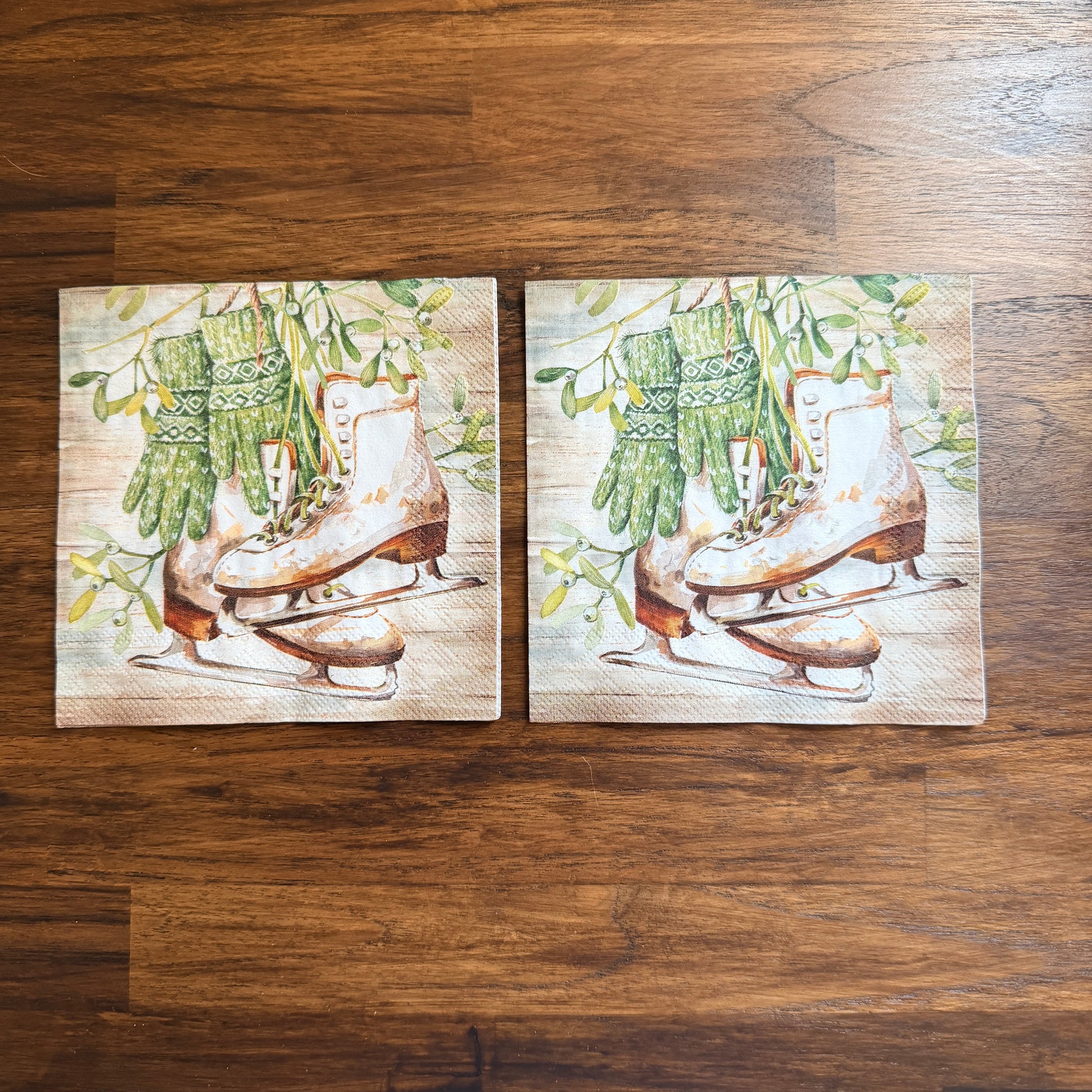 Cocktail size decoupage napkin featuring vintage white leather ice skates with green fair isle mittens hanging from wild mistletoe sprigs on a warm wood grain background set of 2