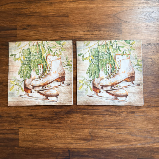 Cocktail size decoupage napkin featuring vintage white leather ice skates with green fair isle mittens hanging from wild mistletoe sprigs on a warm wood grain background set of 2