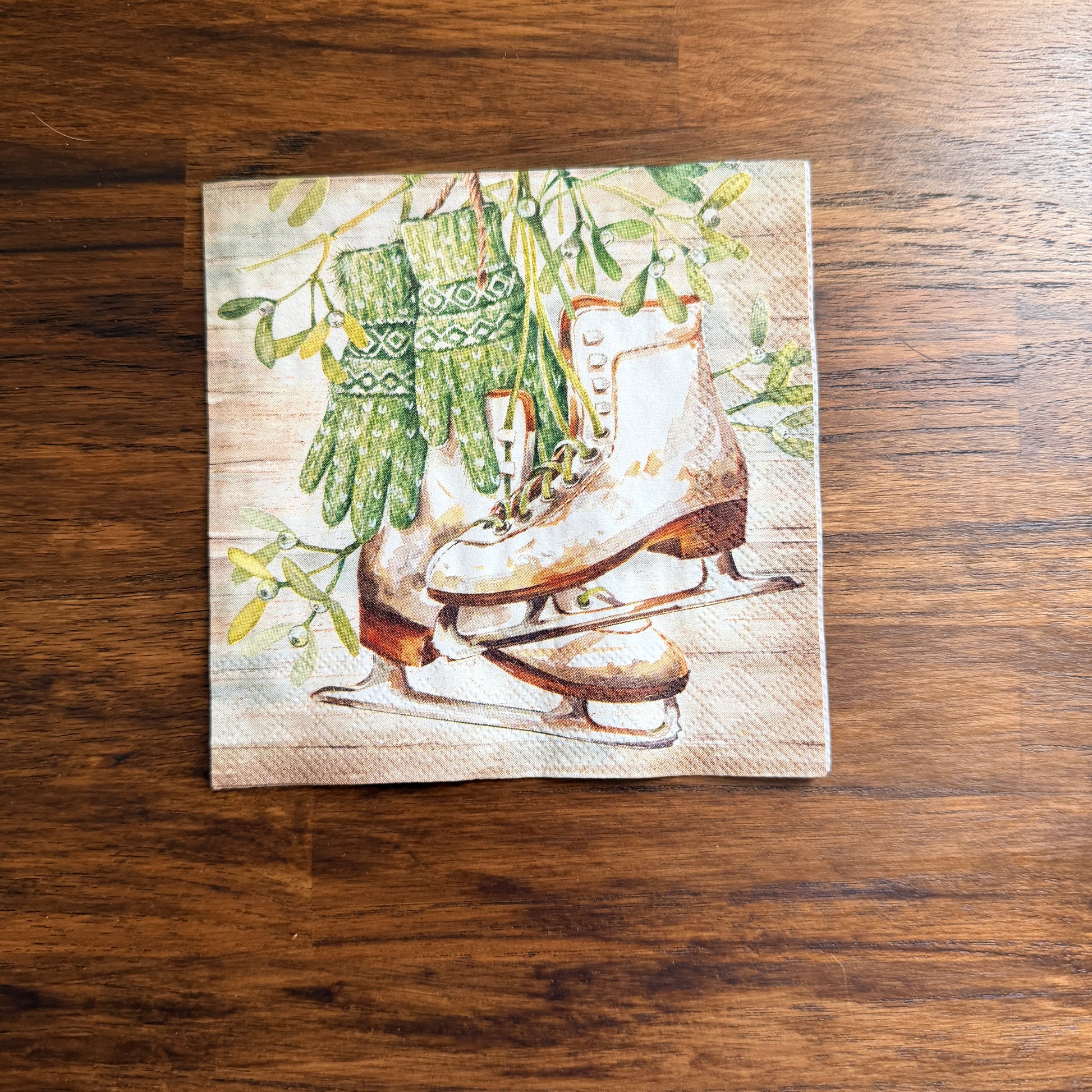 Cocktail size decoupage napkin featuring vintage white leather ice skates with green fair isle mittens hanging from wild mistletoe sprigs on a warm wood grain background set of 2