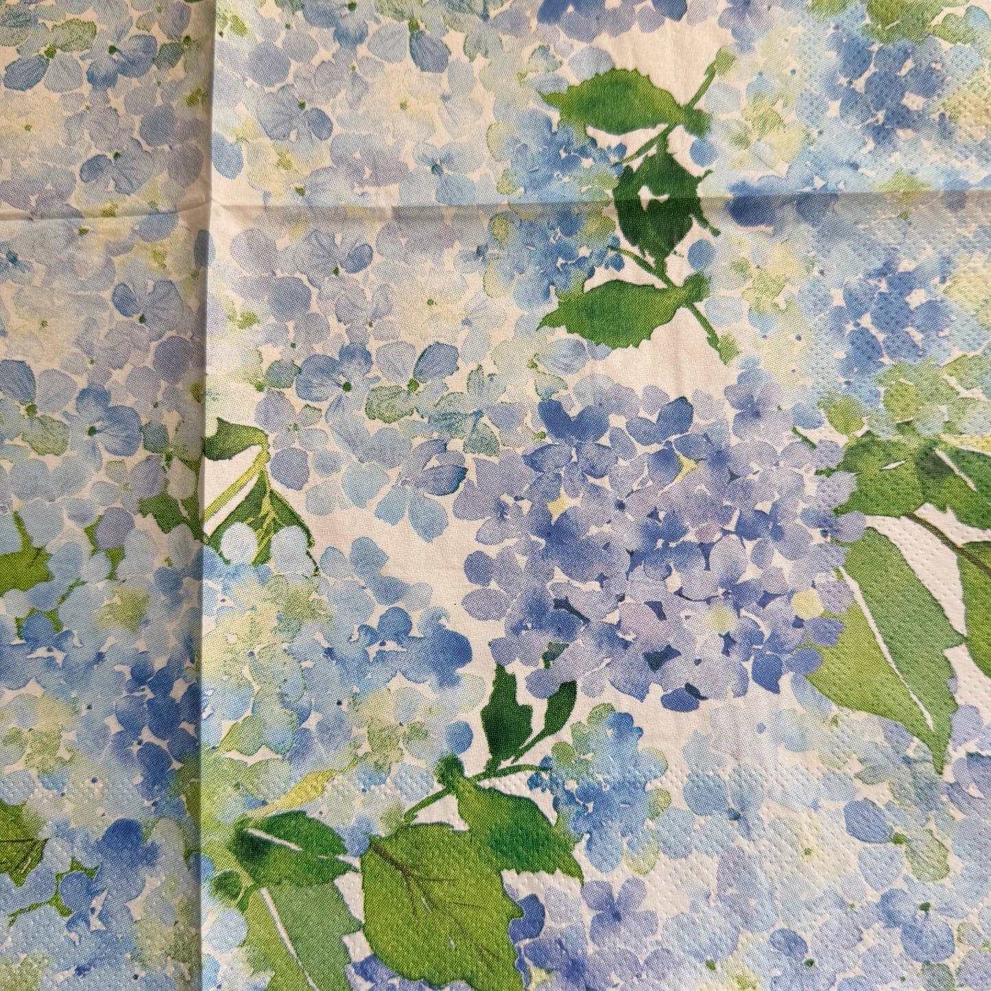 Close up luncheon size decoupage paper napkins with scalloped edges featuring lush watercolor hydrangea clusters in periwinkle blue cornflower and violet with rich green leaves on a creamy white background set of 