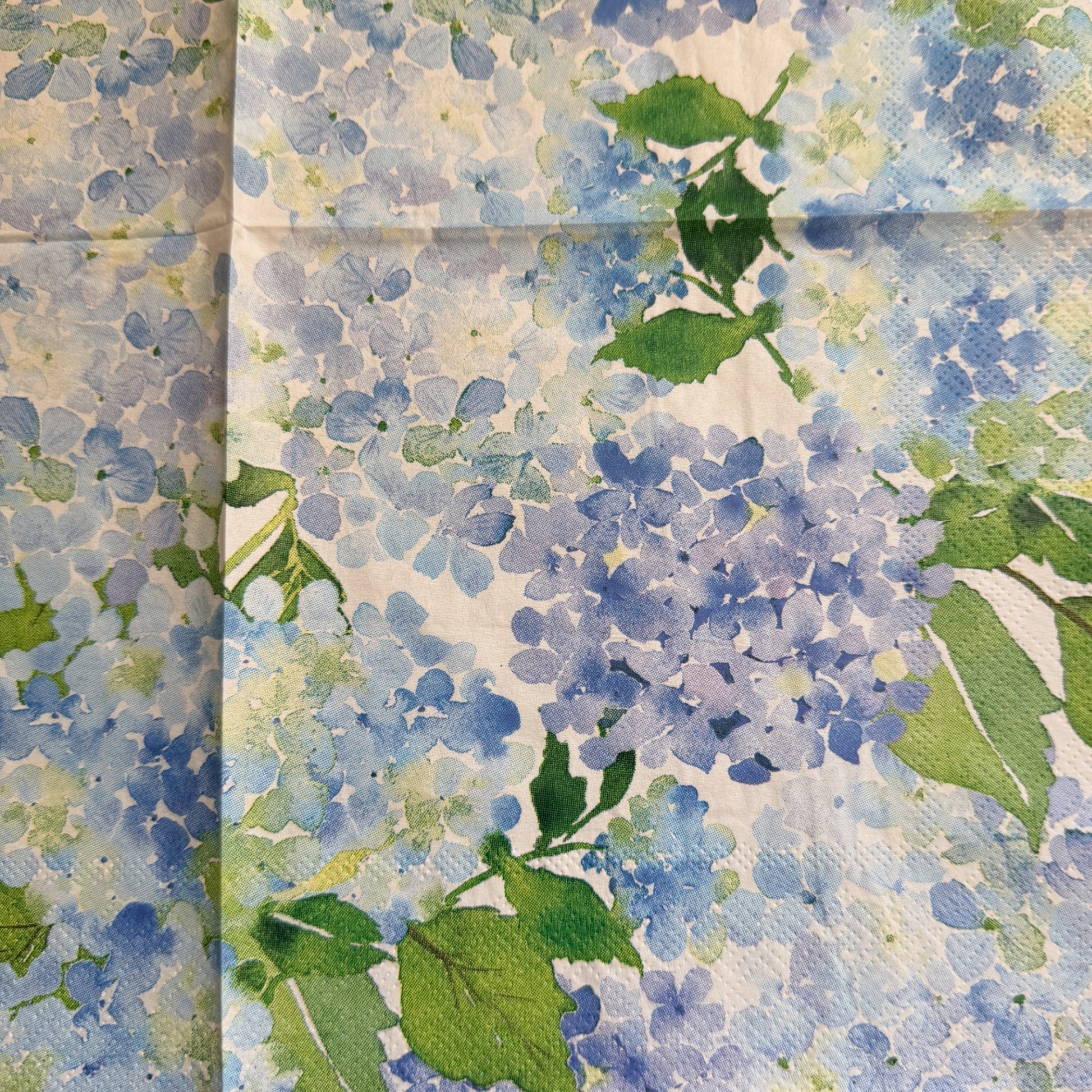 Close up luncheon size decoupage paper napkins with scalloped edges featuring lush watercolor hydrangea clusters in periwinkle blue cornflower and violet with rich green leaves on a creamy white background set of 