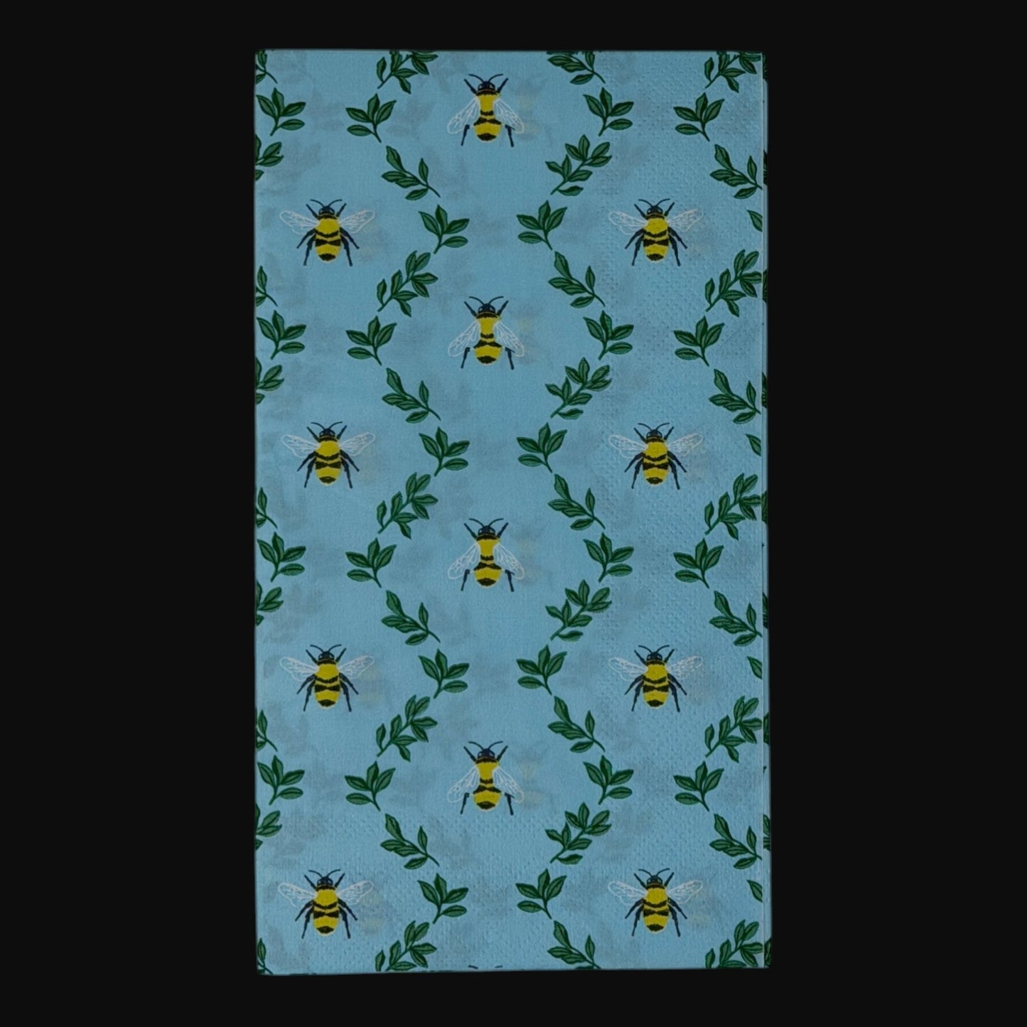  guest-size decoupage paper napkins featuring an all-over print of plump yellow and black bumblebees with white wings scattered among delicate green leaf sprigs on a soft sky blue background with a subtle honeycomb texture, photographed on a black surface.
