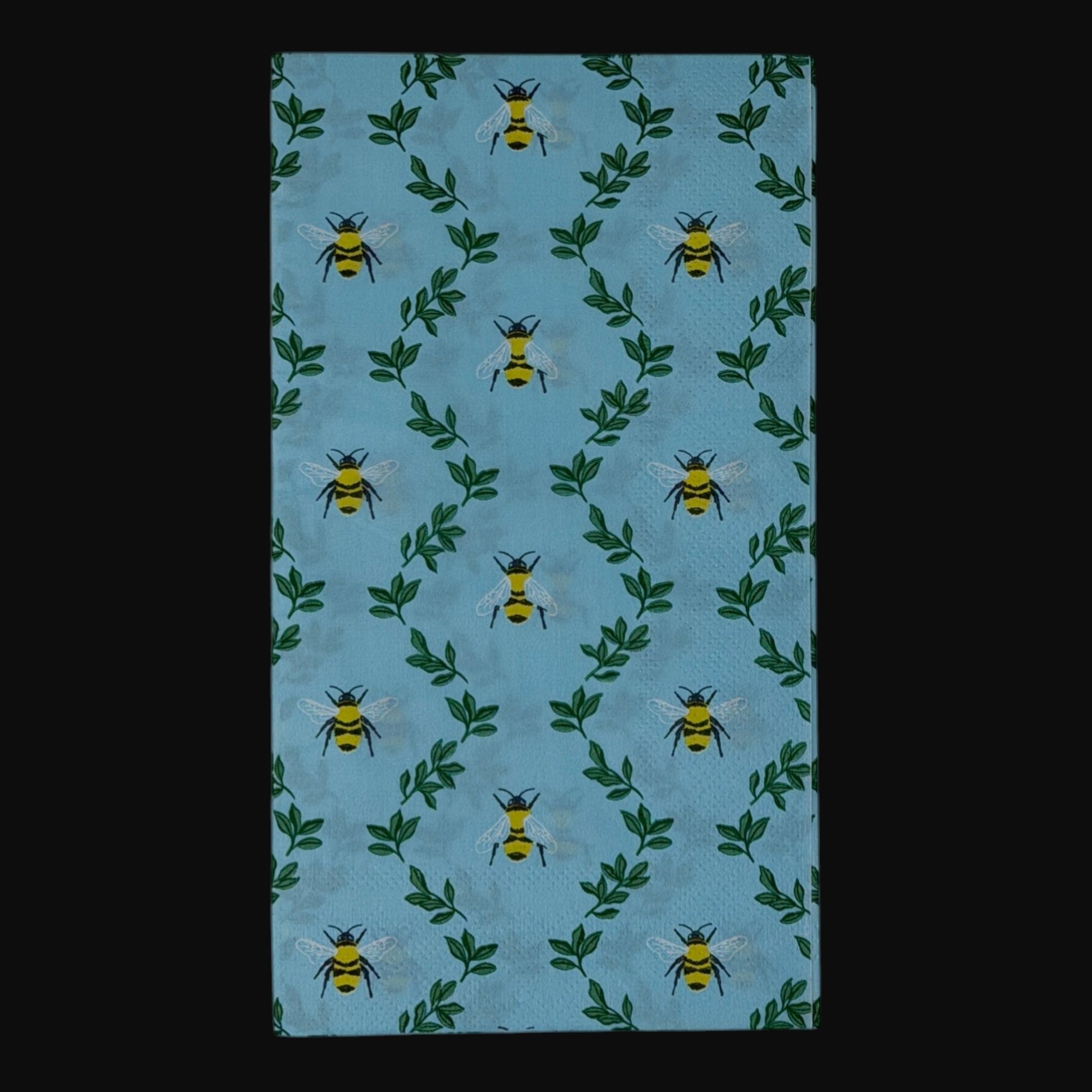  guest-size decoupage paper napkins featuring an all-over print of plump yellow and black bumblebees with white wings scattered among delicate green leaf sprigs on a soft sky blue background with a subtle honeycomb texture, photographed on a black surface.