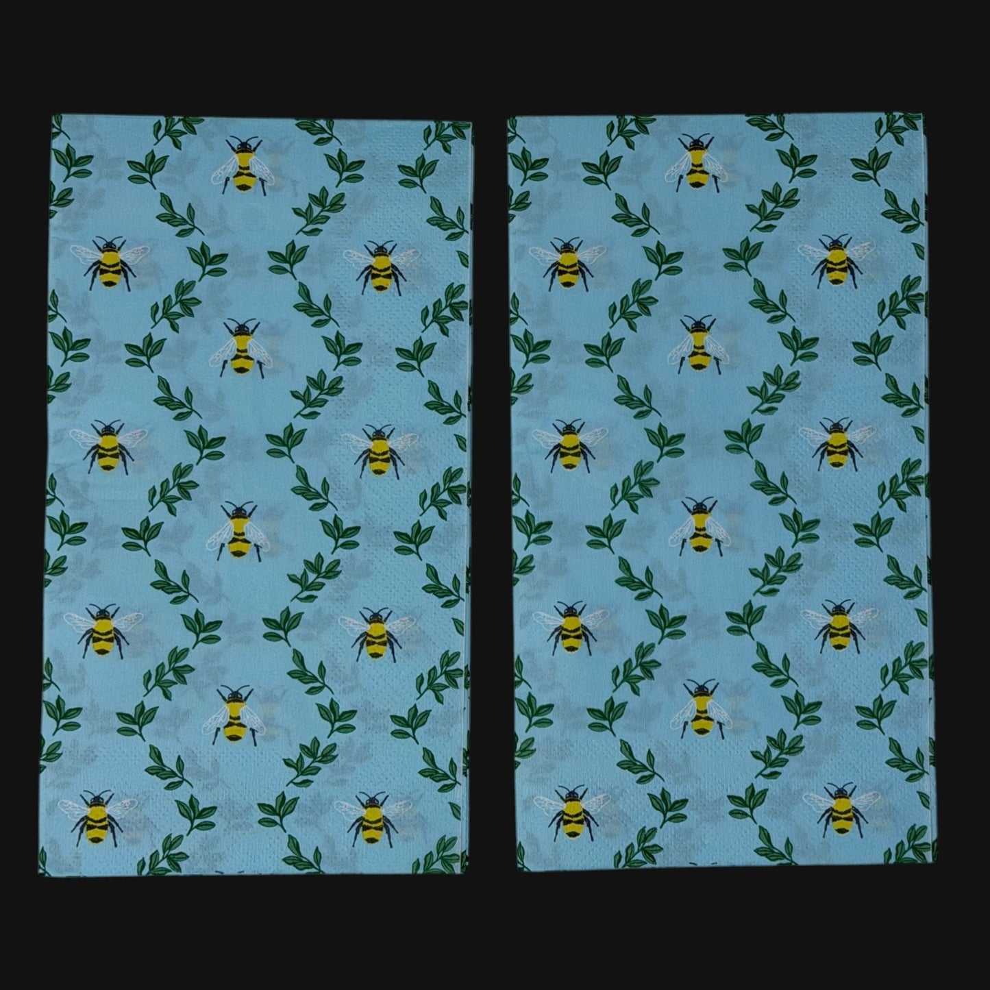 Two guest-size decoupage paper napkins featuring an all-over print of plump yellow and black bumblebees with white wings scattered among delicate green leaf sprigs on a soft sky blue background with a subtle honeycomb texture, photographed on a black surface.