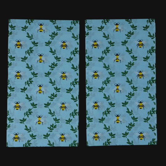 Two guest-size decoupage paper napkins featuring an all-over print of plump yellow and black bumblebees with white wings scattered among delicate green leaf sprigs on a soft sky blue background with a subtle honeycomb texture, photographed on a black surface.