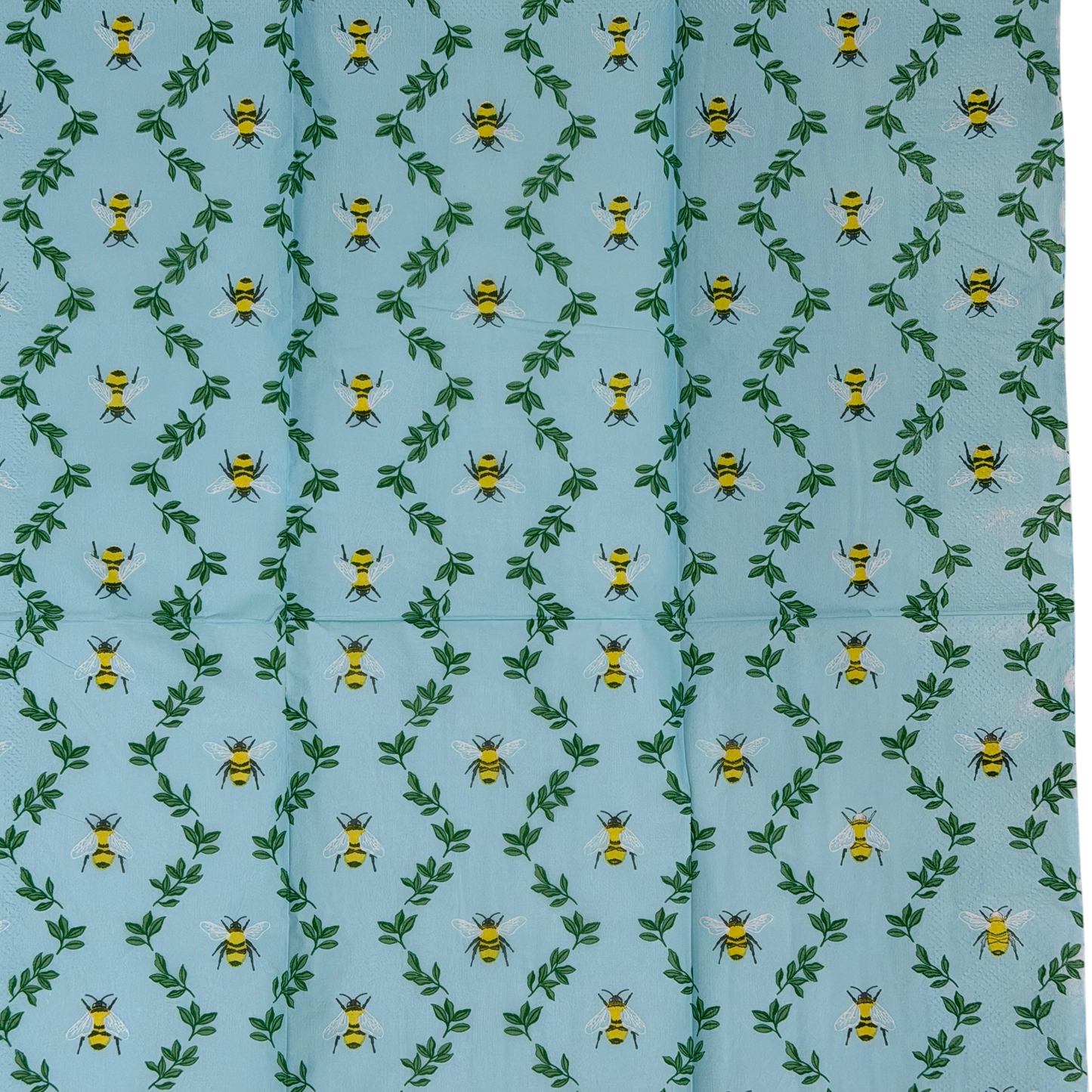 Two guest-size decoupage paper napkins featuring an all-over print of plump yellow and black bumblebees with white wings scattered among delicate green leaf sprigs on a soft sky blue background with a subtle honeycomb texture, photographed on a black surface.