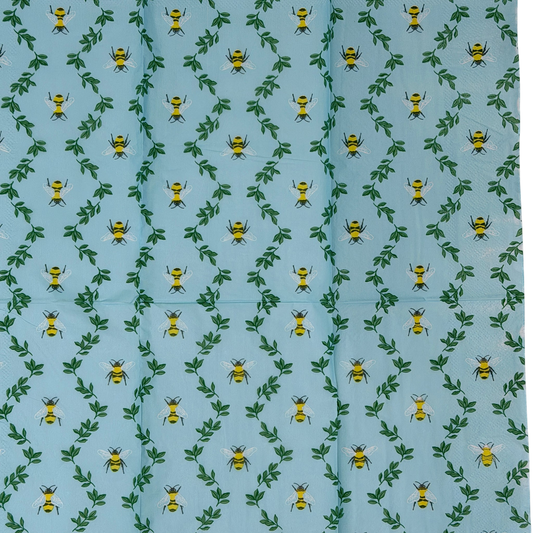 Two guest-size decoupage paper napkins featuring an all-over print of plump yellow and black bumblebees with white wings scattered among delicate green leaf sprigs on a soft sky blue background with a subtle honeycomb texture, photographed on a black surface.