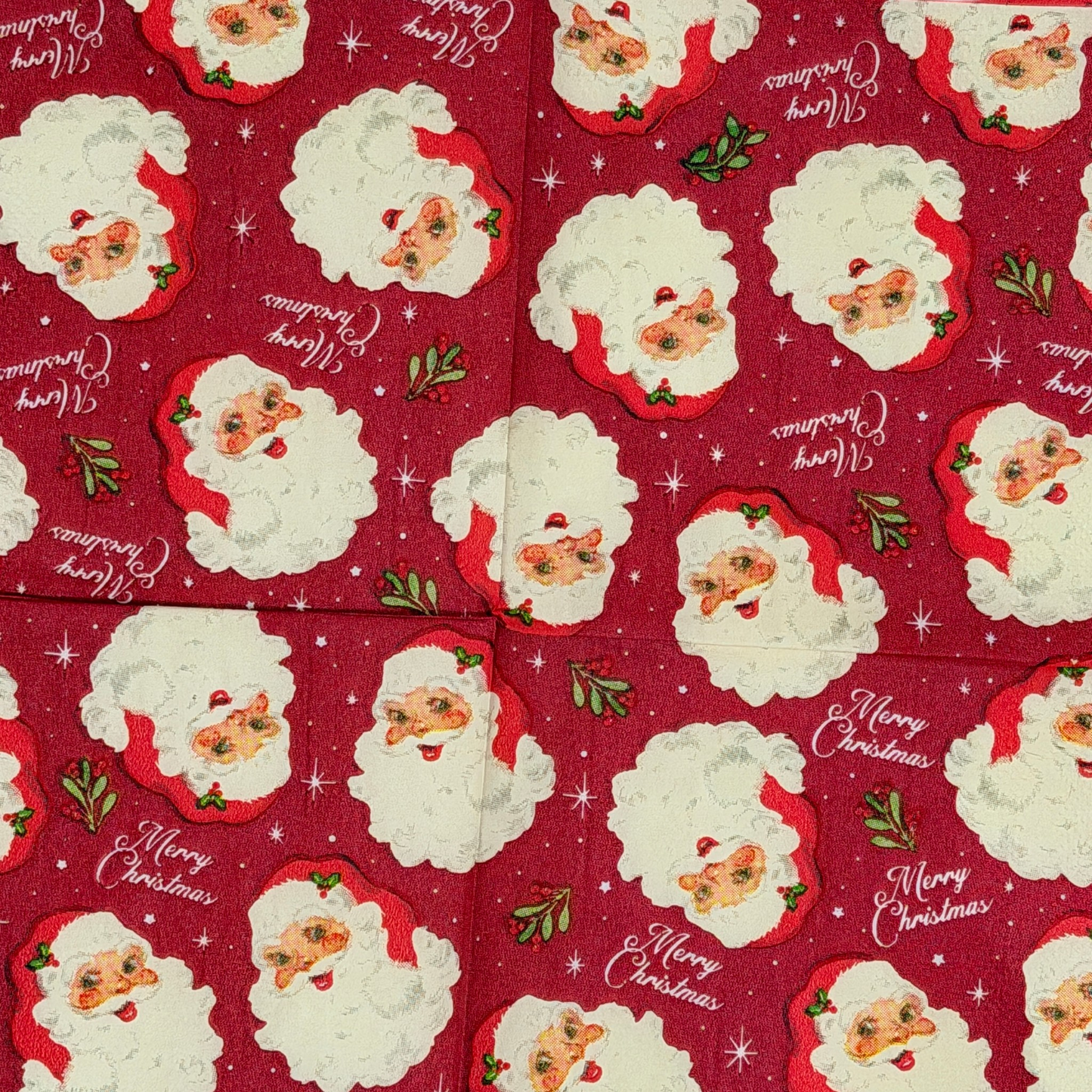 Cocktail size decoupage paper napkin featuring multiple jolly vintage Santa face portraits with fluffy white beards and red hats scattered across a deep crimson background with holly sprigs sparkling stars and Merry Christmas script set of 2