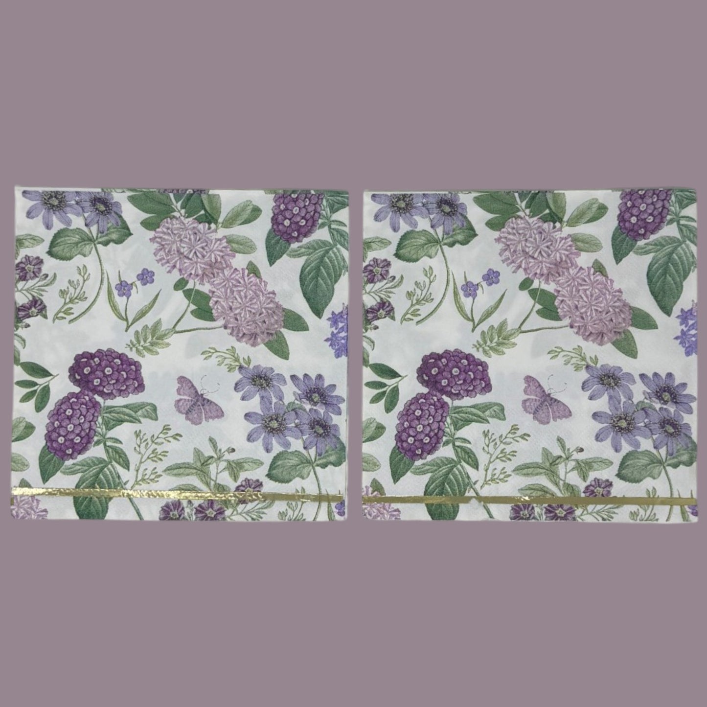 Luncheon-size decoupage paper napkin featuring a vintage botanical illustration print of lavender and purple lilac clusters, periwinkle blue anemones, mauve wildflowers, tiny purple butterflies, and green leaves on a white background with gold foil border stripes, photographed against a white wall.