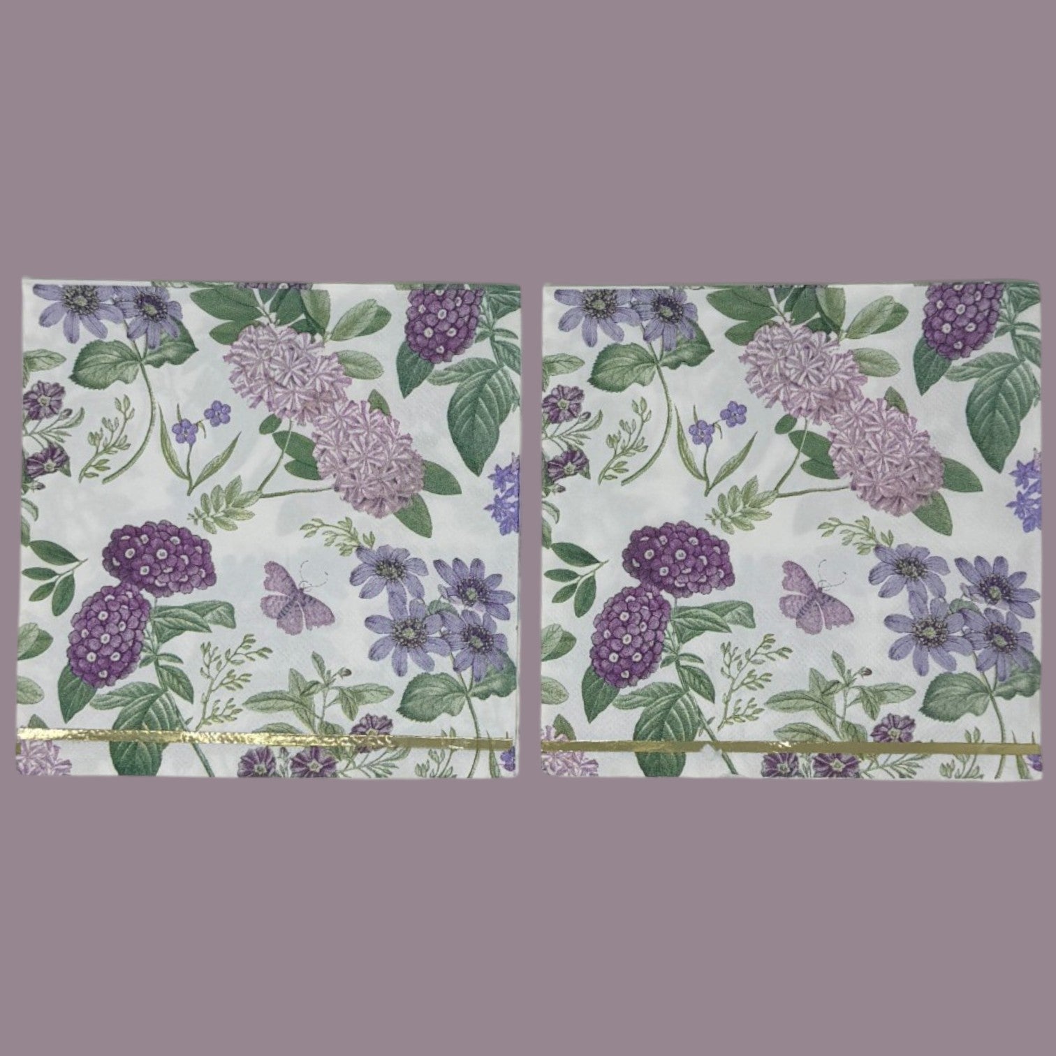 Luncheon-size decoupage paper napkin featuring a vintage botanical illustration print of lavender and purple lilac clusters, periwinkle blue anemones, mauve wildflowers, tiny purple butterflies, and green leaves on a white background with gold foil border stripes, photographed against a white wall.