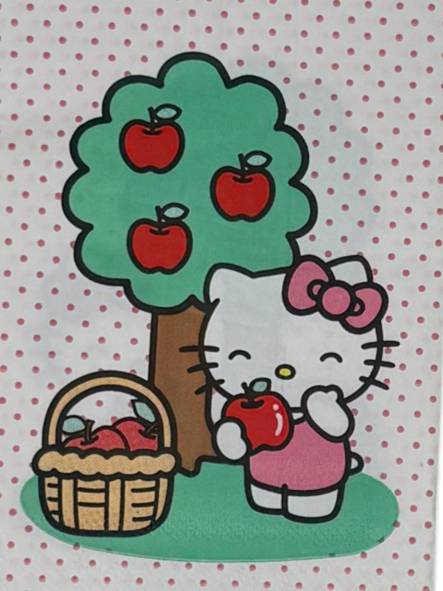 Guest-size decoupage paper napkin featuring a centered illustration of Hello Kitty in a pink bow standing beside a green apple tree with red apples and a wicker basket of apples on a white polka dot background, photographed on a white surface.
