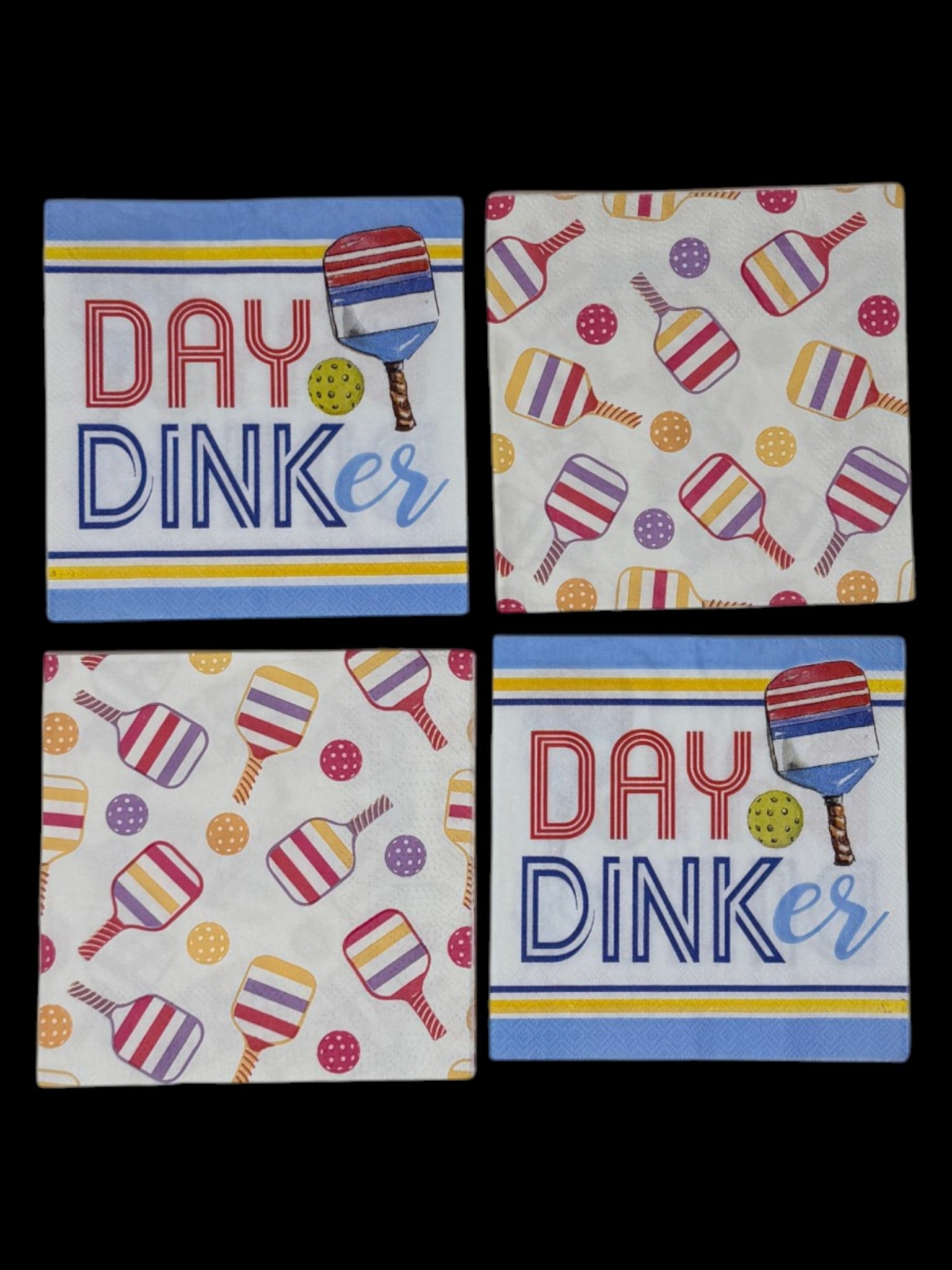 
A bundle of four cocktail-size pickleball themed decoupage paper napkins showing two Day Dinker graphic napkins with bold lettering and a striped paddle on a blue and yellow stripe ground and two colorful all-over tossed paddle and ball pattern napkins on white, photographed on a black surface.