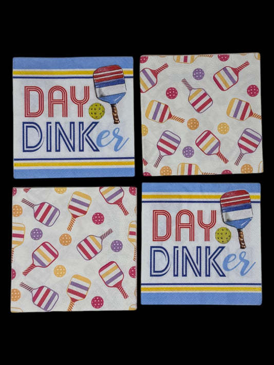 
A bundle of four cocktail-size pickleball themed decoupage paper napkins showing two Day Dinker graphic napkins with bold lettering and a striped paddle on a blue and yellow stripe ground and two colorful all-over tossed paddle and ball pattern napkins on white, photographed on a black surface.