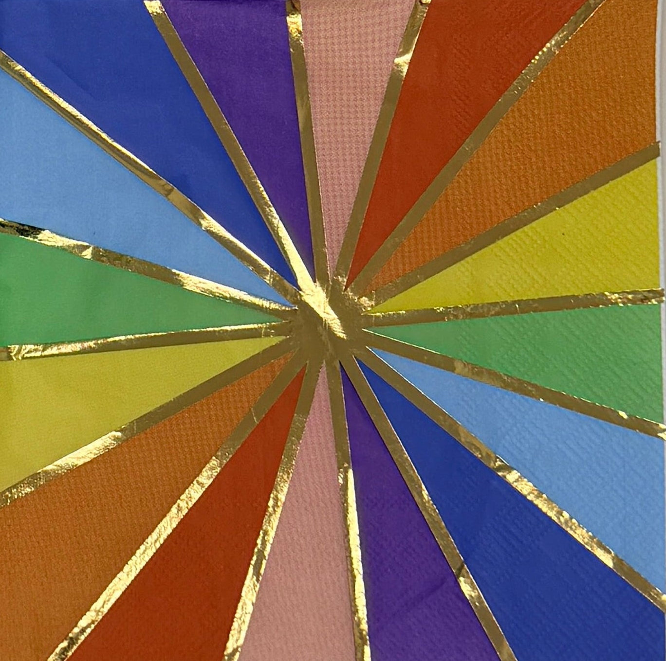 Luncheon-size decoupage paper napkin featuring a bold kaleidoscope pattern of four radiating starburst medallions in cobalt blue, purple, coral, burnt orange, golden yellow, green, and blush pink with crisp white lines and a gold foil starburst accent, photographed unfolded on a light grey surface.