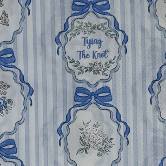 Guest-size decoupage paper napkin featuring a repeating Tying The Knot phrase in elegant script inside an ornate floral cartouche with a blue satin bow on a soft blue and white vertical stripe ground with a scalloped lace border edge, photographed on a black surface.