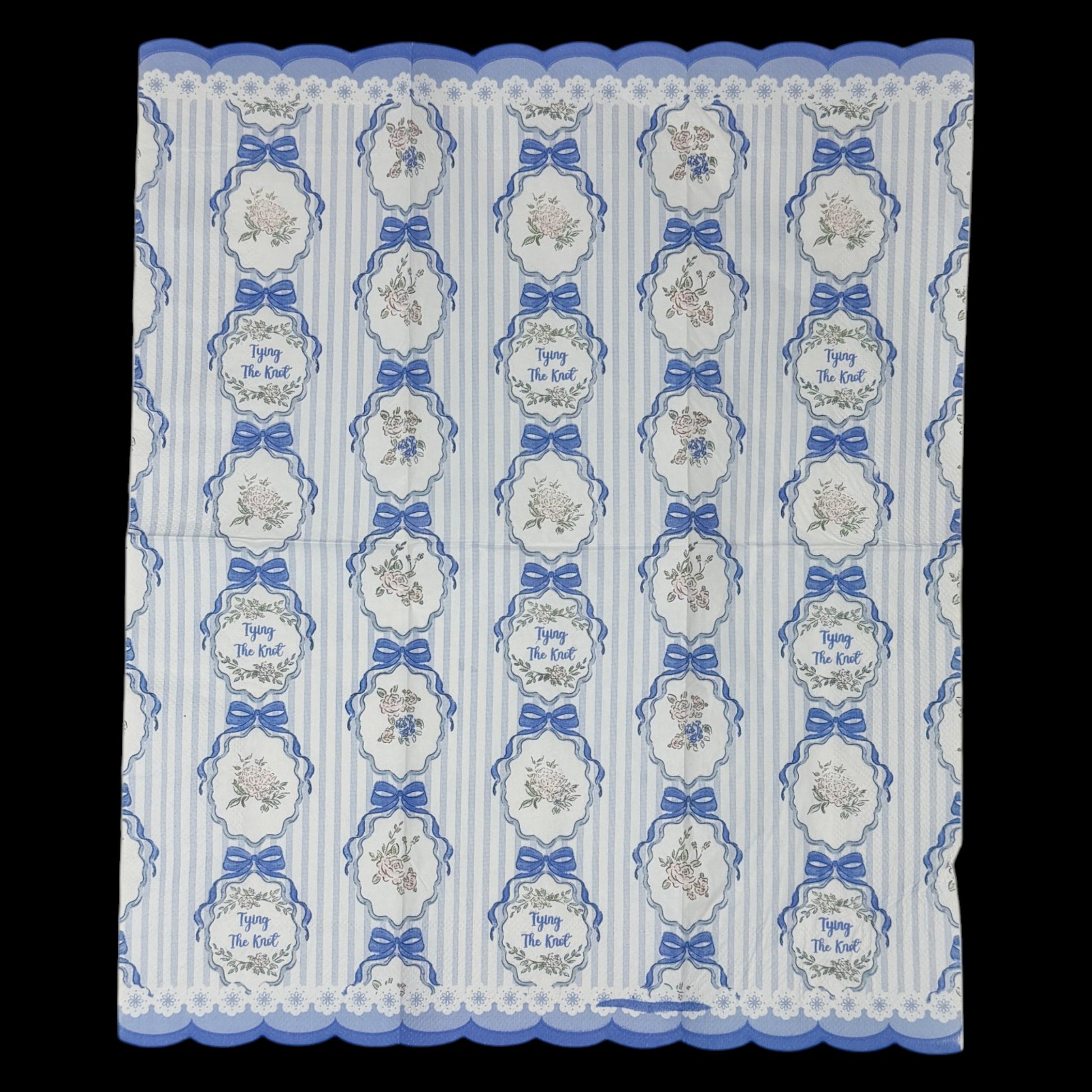 Guest-size decoupage paper napkin featuring a repeating Tying The Knot phrase in elegant script inside an ornate floral cartouche with a blue satin bow on a soft blue and white vertical stripe ground with a scalloped lace border edge, photographed on a black surface.