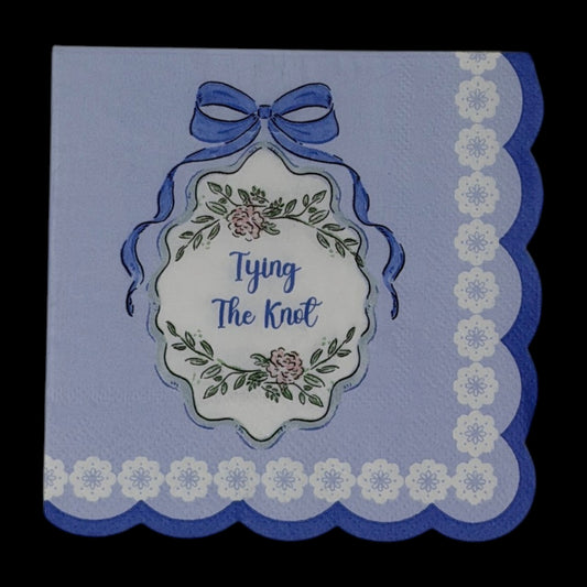 Cocktail-size decoupage paper napkin featuring a centered Tying The Knot medallion with elegant script inside an ornate floral cartouche with a blue satin bow on a solid periwinkle blue ground with a white daisy scalloped lace border edge, photographed on a black surface.
