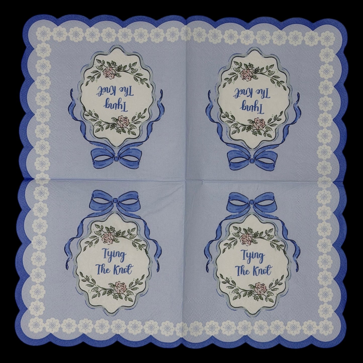 Cocktail-size decoupage paper napkin featuring a centered Tying The Knot medallion with elegant script inside an ornate floral cartouche with a blue satin bow on a solid periwinkle blue ground with a white daisy scalloped lace border edge, photographed on a black surface.