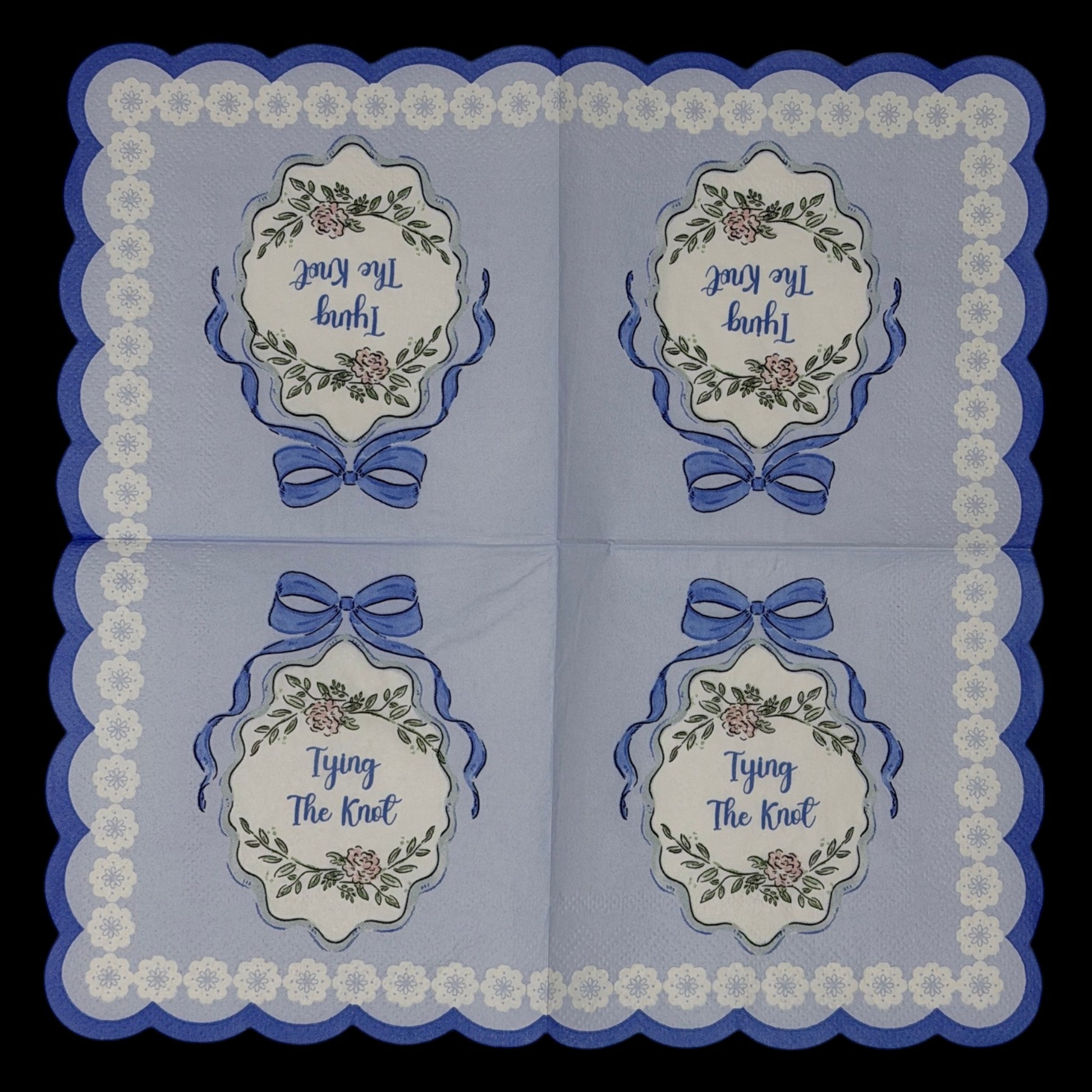 Cocktail-size decoupage paper napkin featuring a centered Tying The Knot medallion with elegant script inside an ornate floral cartouche with a blue satin bow on a solid periwinkle blue ground with a white daisy scalloped lace border edge, photographed on a black surface.
