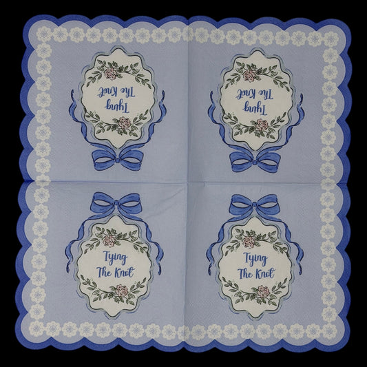 Cocktail-size decoupage paper napkin featuring a centered Tying The Knot medallion with elegant script inside an ornate floral cartouche with a blue satin bow on a solid periwinkle blue ground with a white daisy scalloped lace border edge, photographed on a black surface.