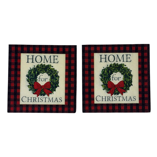 Cocktail-size decoupage paper napkin featuring the phrase Home for Christmas in dark green serif lettering surrounding a lush green boxwood wreath with a full red satin bow on a soft cream ground framed by a deep red and black buffalo check plaid border, photographed on a white surface.