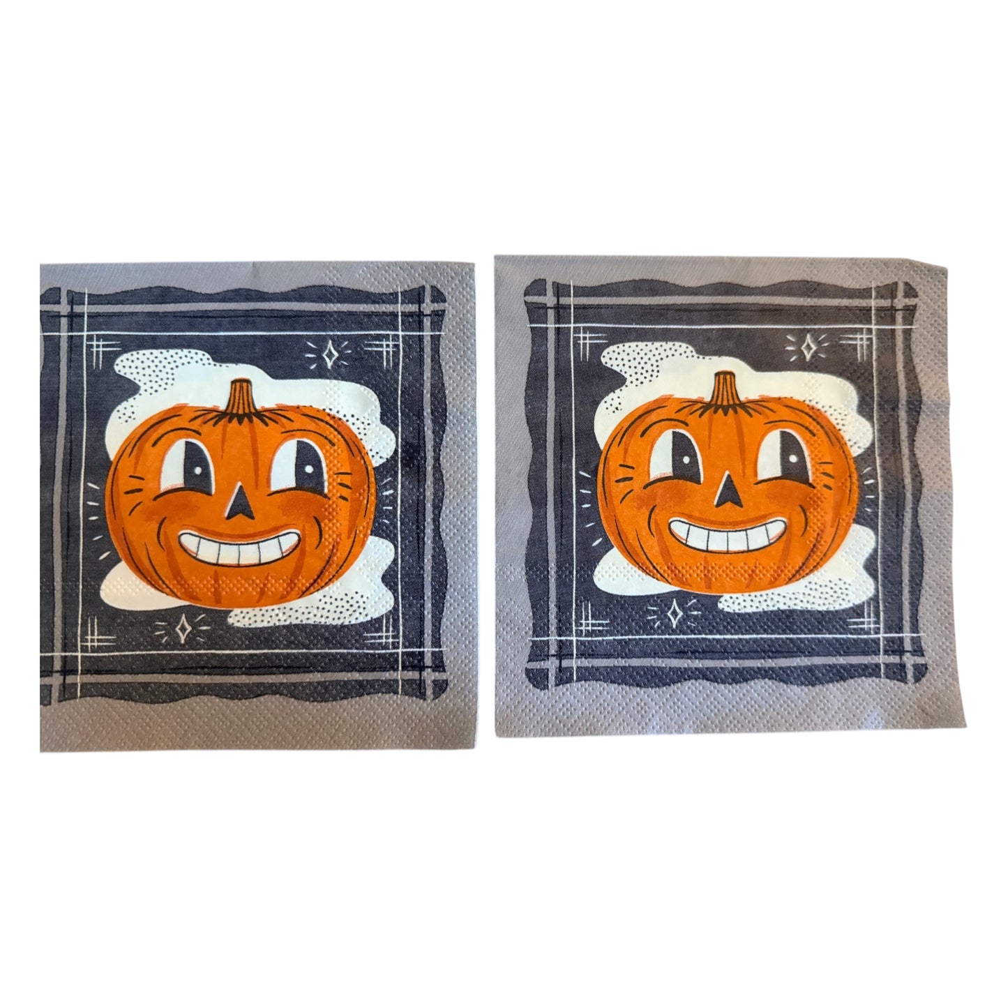 Cocktail-size decoupage paper napkin featuring a bold retro illustration of a grinning cartoon jack-o-lantern with wide eyes and a toothy smile set against a misty white cloud on a deep navy black background with a graphic illustrated border and diamond star accents, photographed on a warm wood surface.