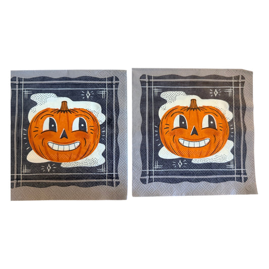 Cocktail-size decoupage paper napkin featuring a bold retro illustration of a grinning cartoon jack-o-lantern with wide eyes and a toothy smile set against a misty white cloud on a deep navy black background with a graphic illustrated border and diamond star accents, photographed on a warm wood surface.