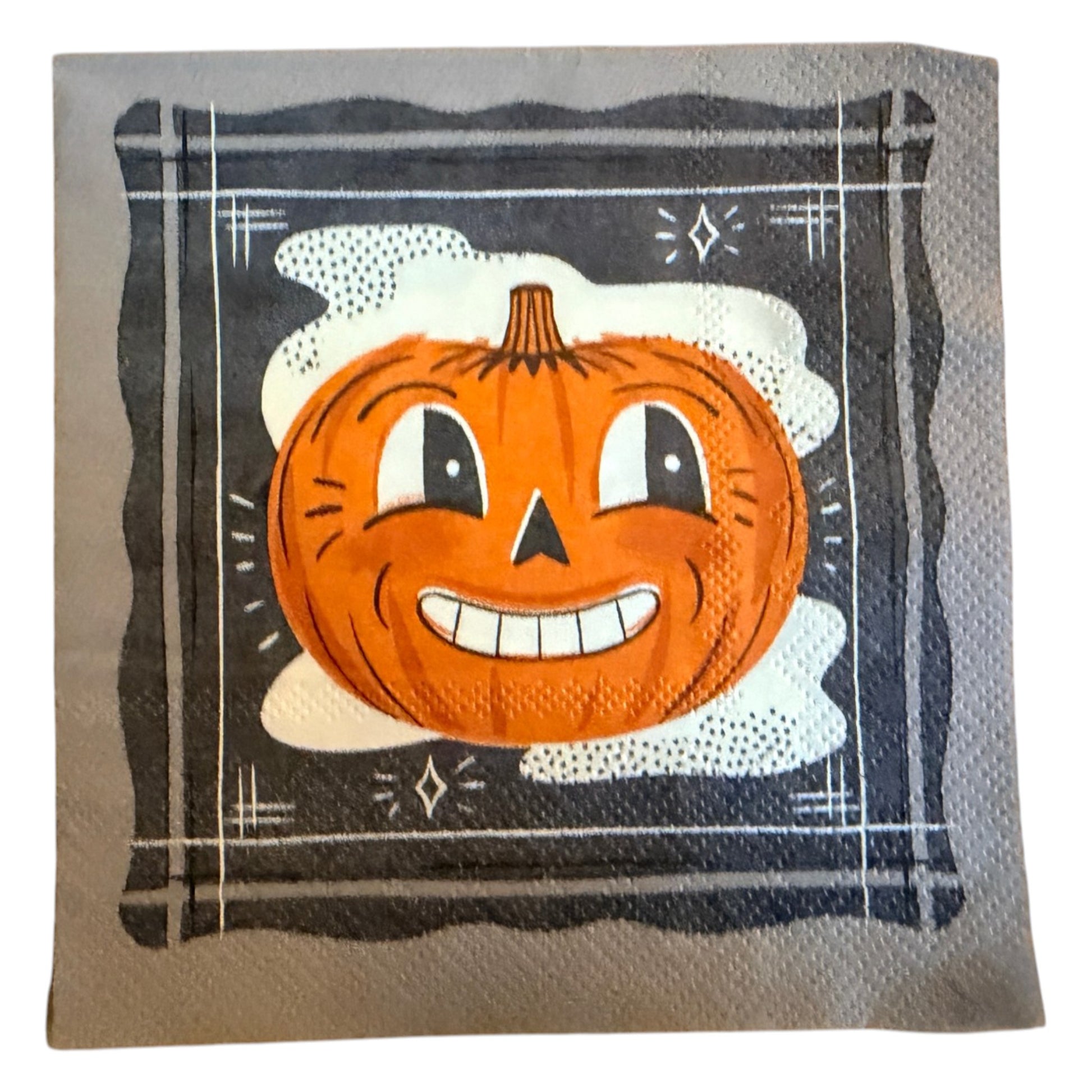 Cocktail-size decoupage paper napkin featuring a bold retro illustration of a grinning cartoon jack-o-lantern with wide eyes and a toothy smile set against a misty white cloud on a deep navy black background with a graphic illustrated border and diamond star accents, photographed on a warm wood surface.