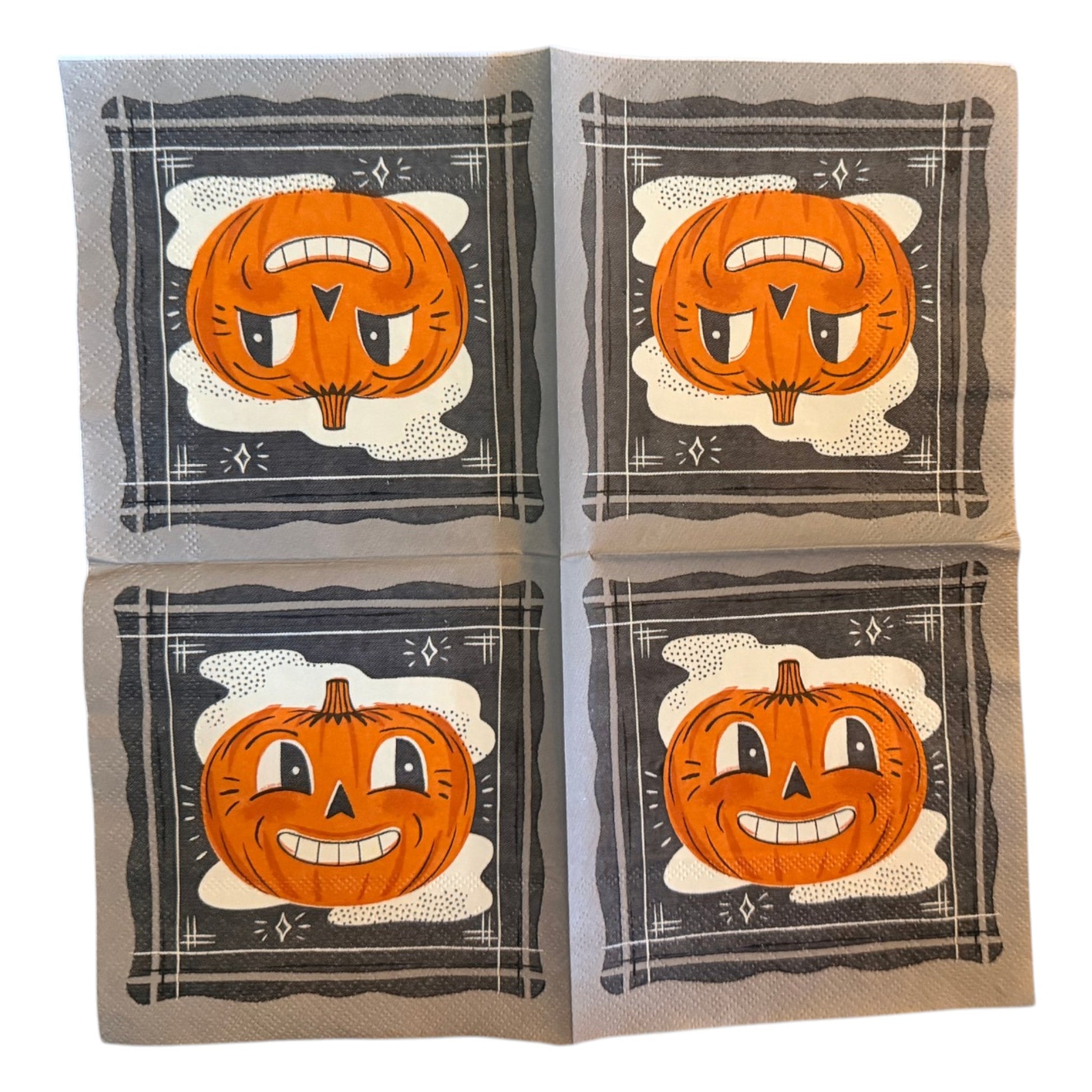 Cocktail-size decoupage paper napkin featuring a bold retro illustration of a grinning cartoon jack-o-lantern with wide eyes and a toothy smile set against a misty white cloud on a deep navy black background with a graphic illustrated border and diamond star accents, photographed on a warm wood surface.