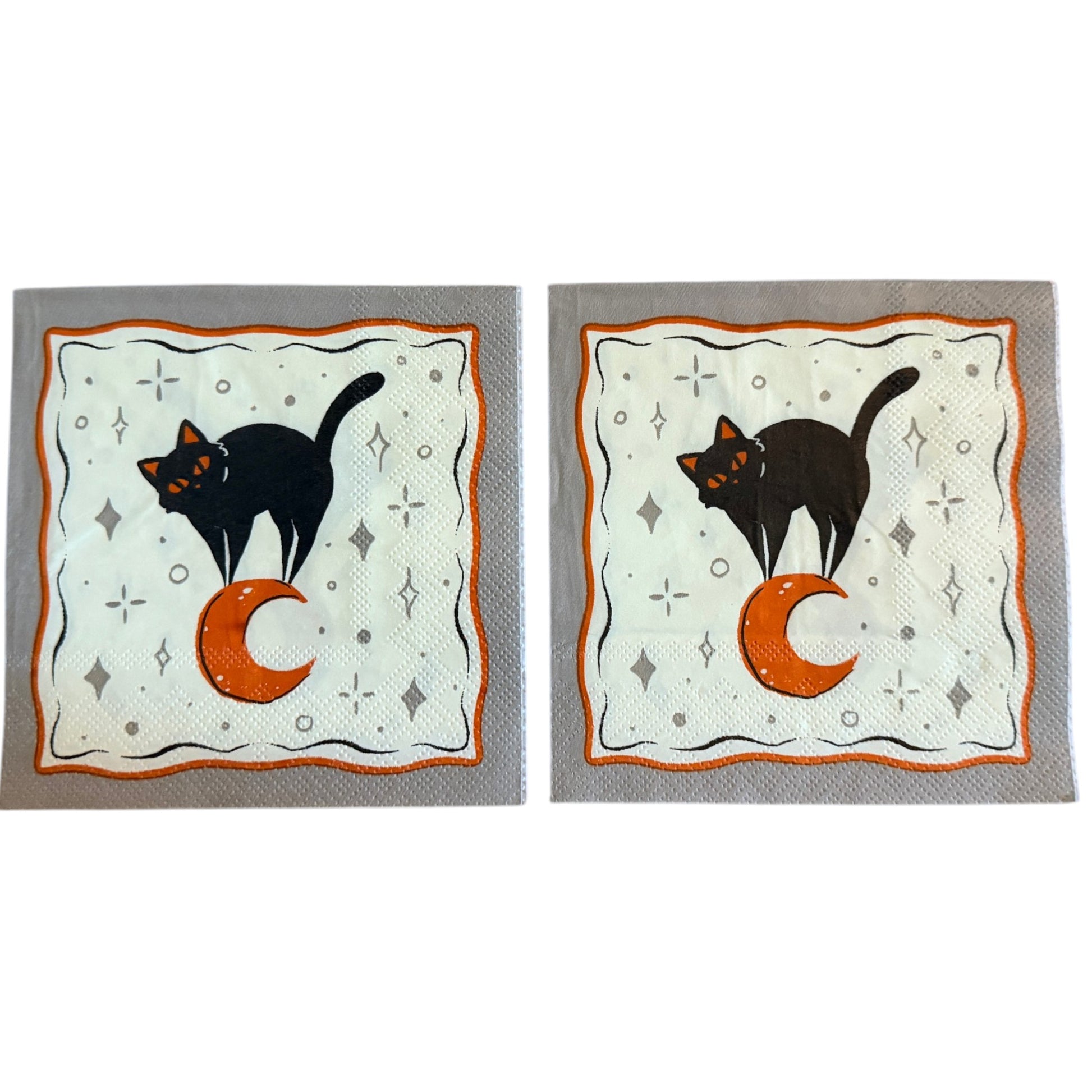 Cocktail-size decoupage paper napkin featuring a sleek black cat with glowing orange eyes sitting beside a large orange crescent moon on a soft white background scattered with grey star and diamond accents framed by a wavy orange border on grey, photographed on a warm wood surface.