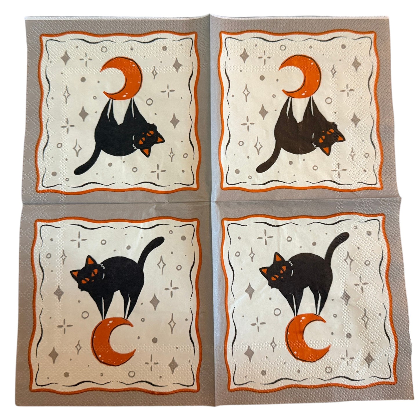 Cocktail-size decoupage paper napkin featuring a sleek black cat with glowing orange eyes sitting beside a large orange crescent moon on a soft white background scattered with grey star and diamond accents framed by a wavy orange border on grey, photographed on a warm wood surface.