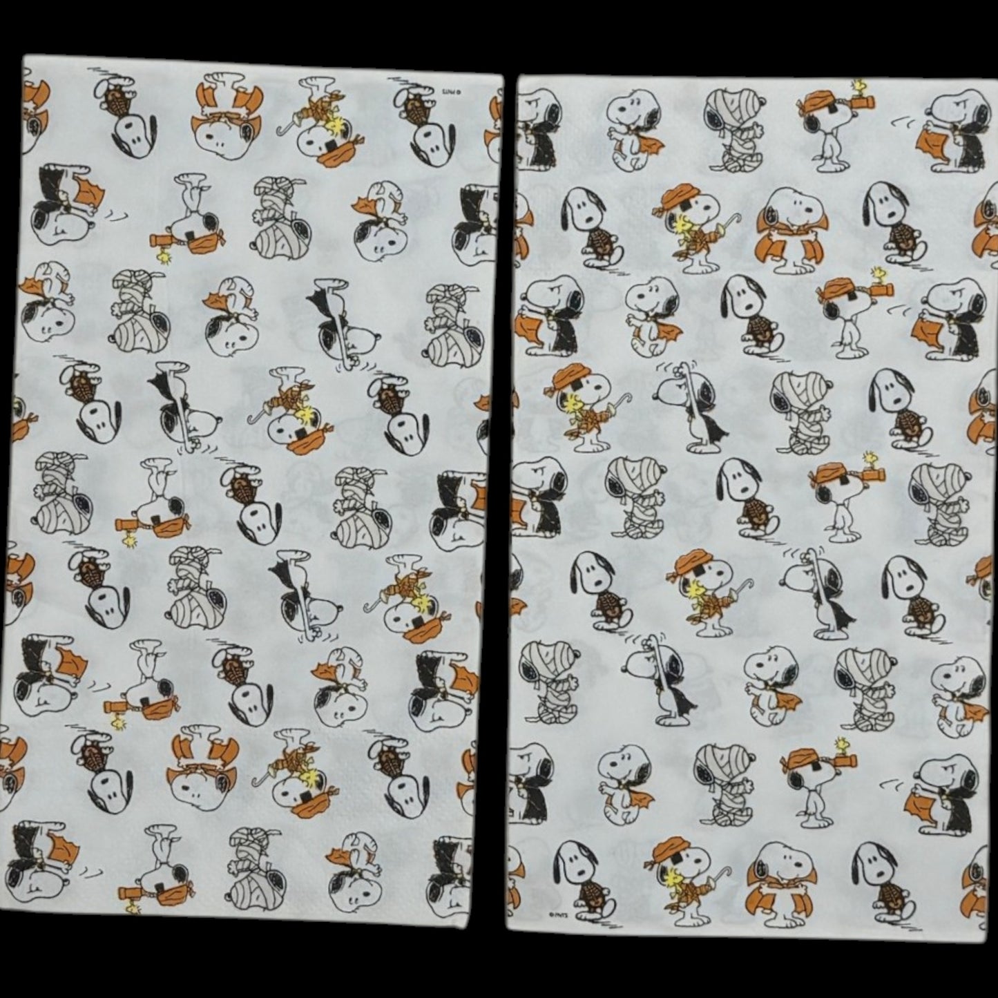 Guest-size decoupage paper napkin featuring an all-over tossed pattern of Snoopy and Woodstock in various Halloween costumes including vampire mummy and witch in classic Peanuts comic illustration style on a soft white background, photographed on a black surface.