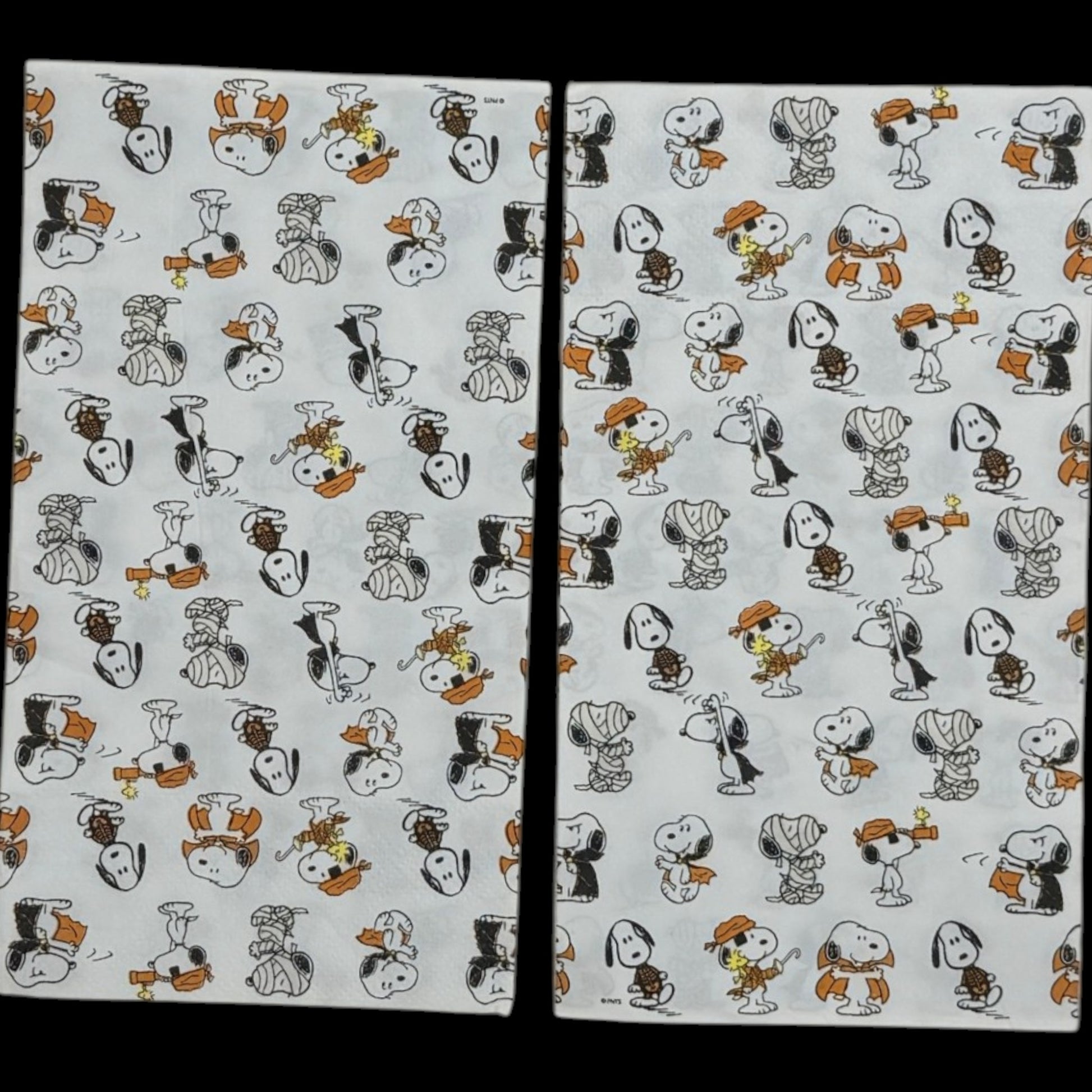 Guest-size decoupage paper napkin featuring an all-over tossed pattern of Snoopy and Woodstock in various Halloween costumes including vampire mummy and witch in classic Peanuts comic illustration style on a soft white background, photographed on a black surface.