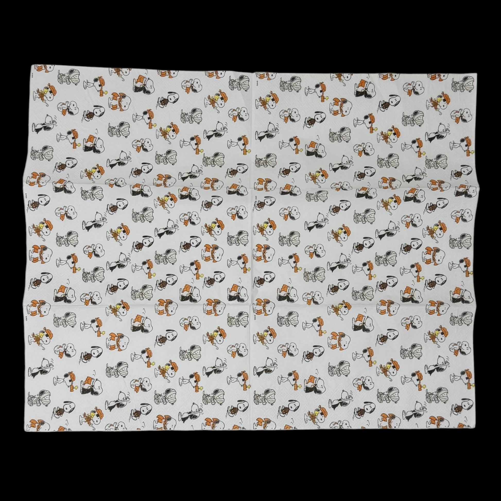 Guest-size decoupage paper napkin featuring an all-over tossed pattern of Snoopy and Woodstock in various Halloween costumes including vampire mummy and witch in classic Peanuts comic illustration style on a soft white background, photographed on a black surface.