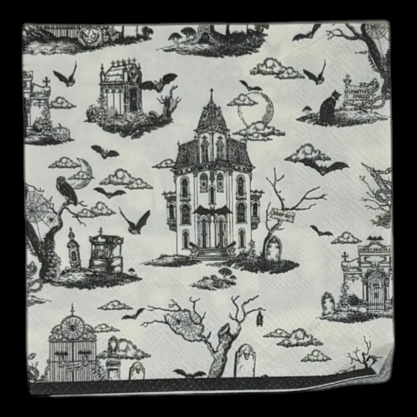 Cocktail-size decoupage paper napkin featuring a richly detailed all-over Halloween toile print in black ink on soft cream white showing a Victorian haunted mansion flying bats crescent moon owl cemetery gates tombstones a black cat in a top hat gnarled dead trees and a zombie hand rising from the earth, photographed on a black surface.