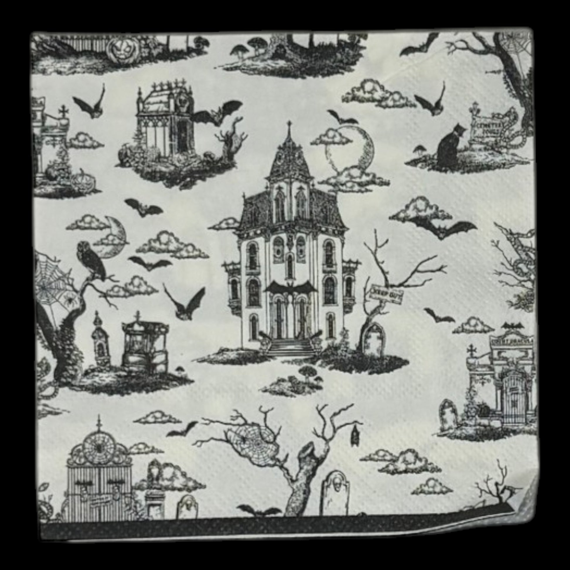 Cocktail-size decoupage paper napkin featuring a richly detailed all-over Halloween toile print in black ink on soft cream white showing a Victorian haunted mansion flying bats crescent moon owl cemetery gates tombstones a black cat in a top hat gnarled dead trees and a zombie hand rising from the earth, photographed on a black surface.
