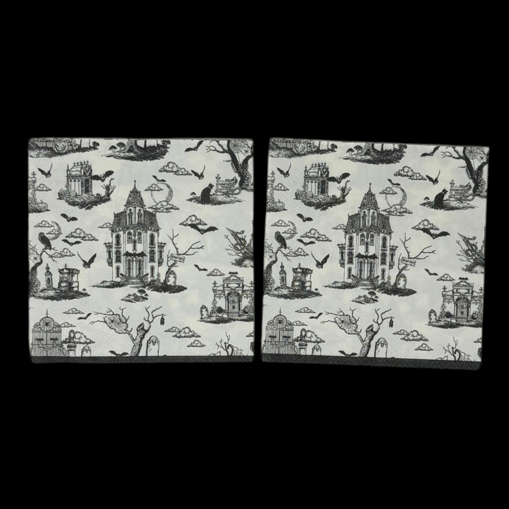 Cocktail-size decoupage paper napkin featuring a richly detailed all-over Halloween toile print in black ink on soft cream white showing a Victorian haunted mansion flying bats crescent moon owl cemetery gates tombstones a black cat in a top hat gnarled dead trees and a zombie hand rising from the earth, photographed on a black surface.
