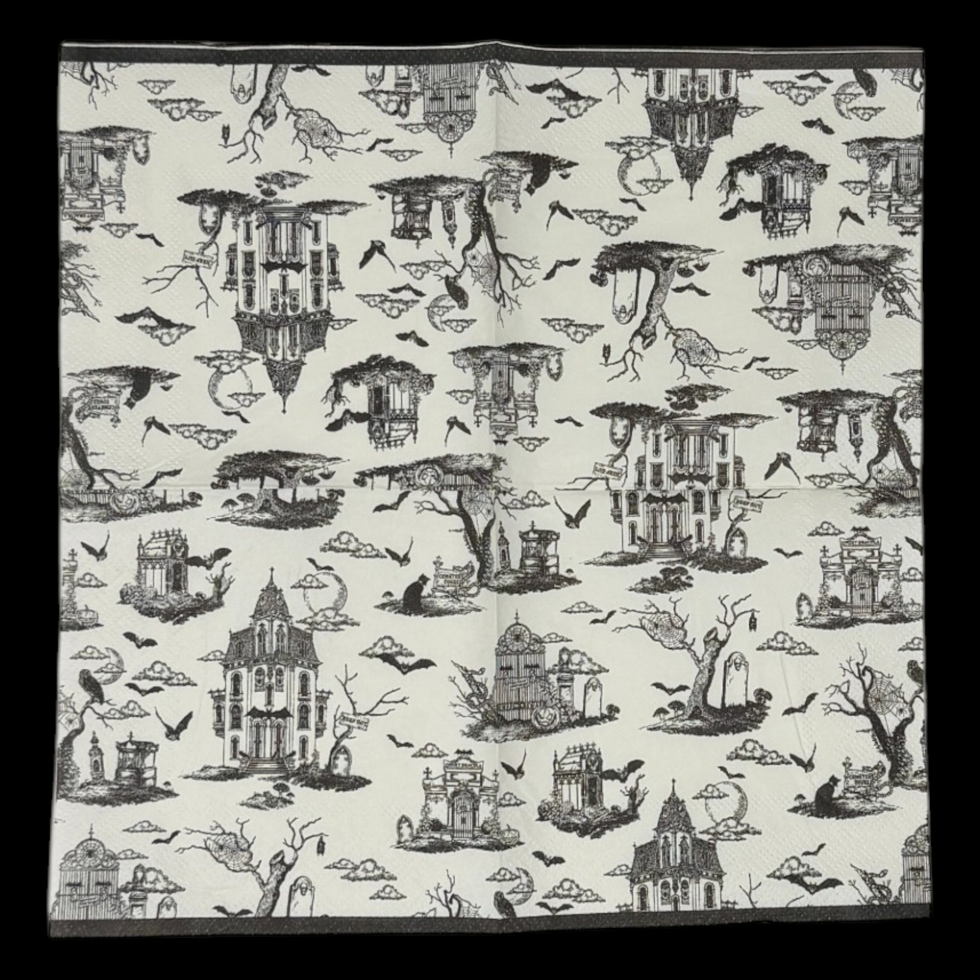 Cocktail-size decoupage paper napkin featuring a richly detailed all-over Halloween toile print in black ink on soft cream white showing a Victorian haunted mansion flying bats crescent moon owl cemetery gates tombstones a black cat in a top hat gnarled dead trees and a zombie hand rising from the earth, photographed on a black surface.
