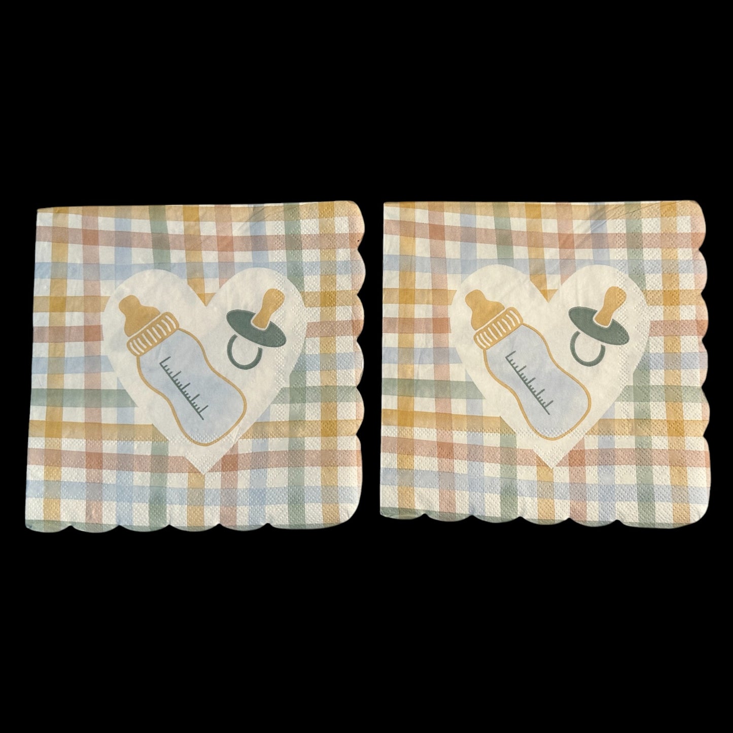 Close-up of two scalloped-edge luncheon napkins featuring a pastel gingham pattern in yellow, peach, blue, and green with a white heart in the center displaying a baby bottle and pacifier design.