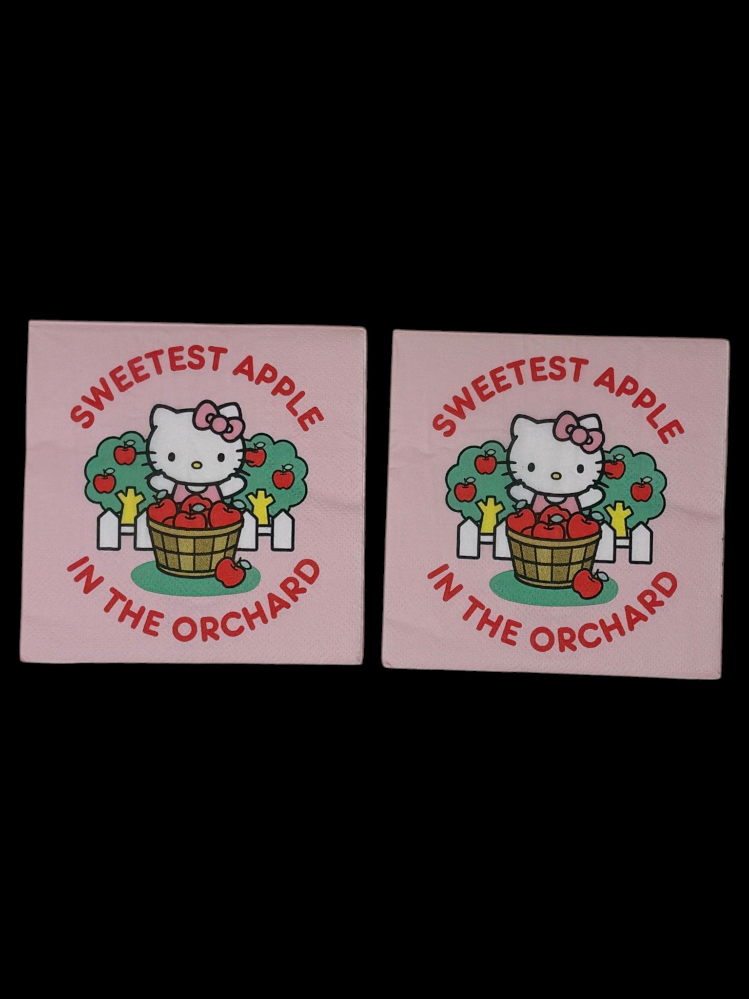 Cocktail-size decoupage paper napkin featuring Hello Kitty standing behind a wooden barrel of red apples with green apple trees and a white picket fence, surrounded by the phrase Sweetest Apple in the Orchard in bold red lettering on a soft blush pink background.