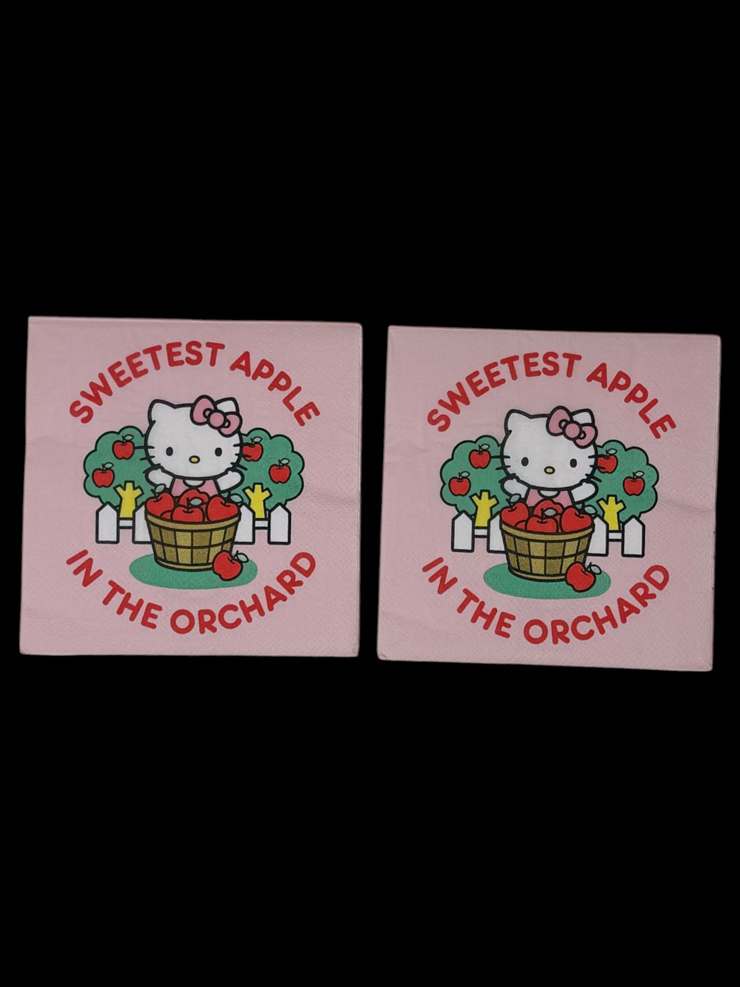 Cocktail-size decoupage paper napkin featuring Hello Kitty standing behind a wooden barrel of red apples with green apple trees and a white picket fence, surrounded by the phrase Sweetest Apple in the Orchard in bold red lettering on a soft blush pink background.