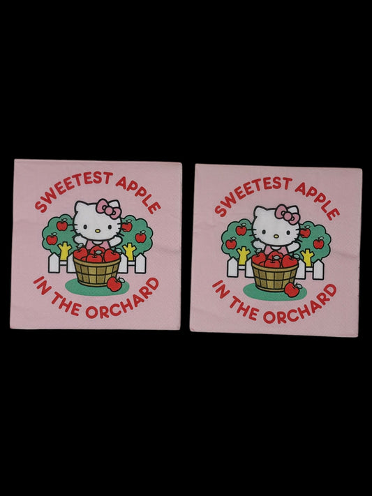 Cocktail-size decoupage paper napkin featuring Hello Kitty standing behind a wooden barrel of red apples with green apple trees and a white picket fence, surrounded by the phrase Sweetest Apple in the Orchard in bold red lettering on a soft blush pink background.