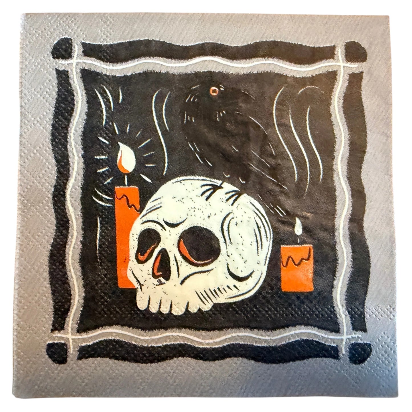 A square paper napkin featuring a gothic illustration of a skull, a raven, and two orange candles on a black background with a white border.