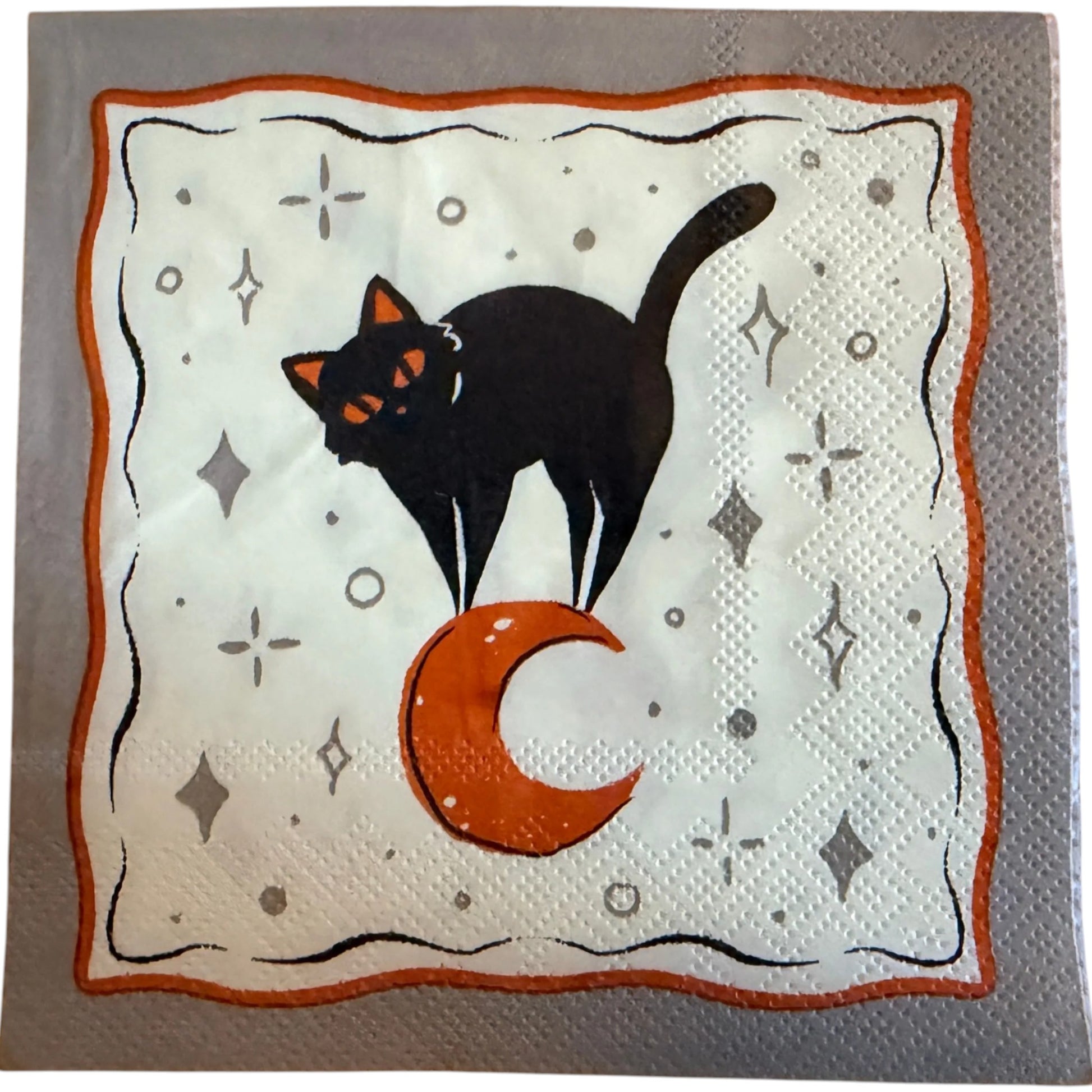 Cocktail-size decoupage paper napkin featuring a sleek black cat with glowing orange eyes sitting beside a large orange crescent moon on a soft white background scattered with grey star and diamond accents framed by a wavy orange border on grey, photographed on a warm wood surface.