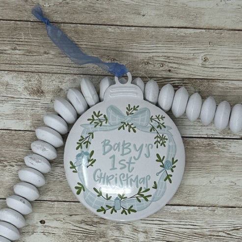Decorative blue and white metal ornament for baby's first Christmas on a wooden surface.  Features a blue sheer ribbon for hanging