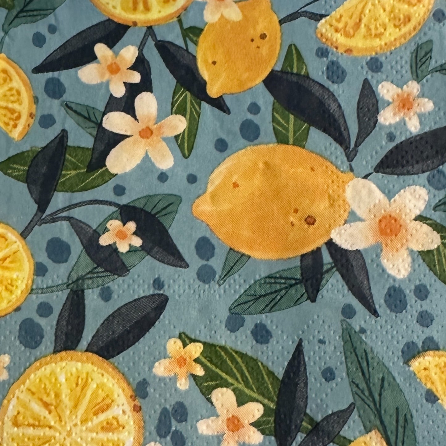 Cocktail-size decoupage paper napkin featuring a lush all-over botanical print of golden whole lemons and halved citrus slices with white orange blossom flowers and rich dark and forest green leaves on a soft dusty blue background with subtle dot accents, photographed on a black surface.