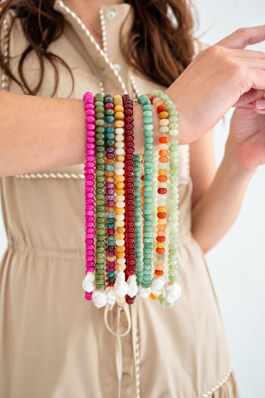 A close-up of a person wearing a beige dress, showing several colorful beaded stretch bracelets on their wrist. The bracelets feature various bead patterns and colors including pink, green, orange, yellow, and white.